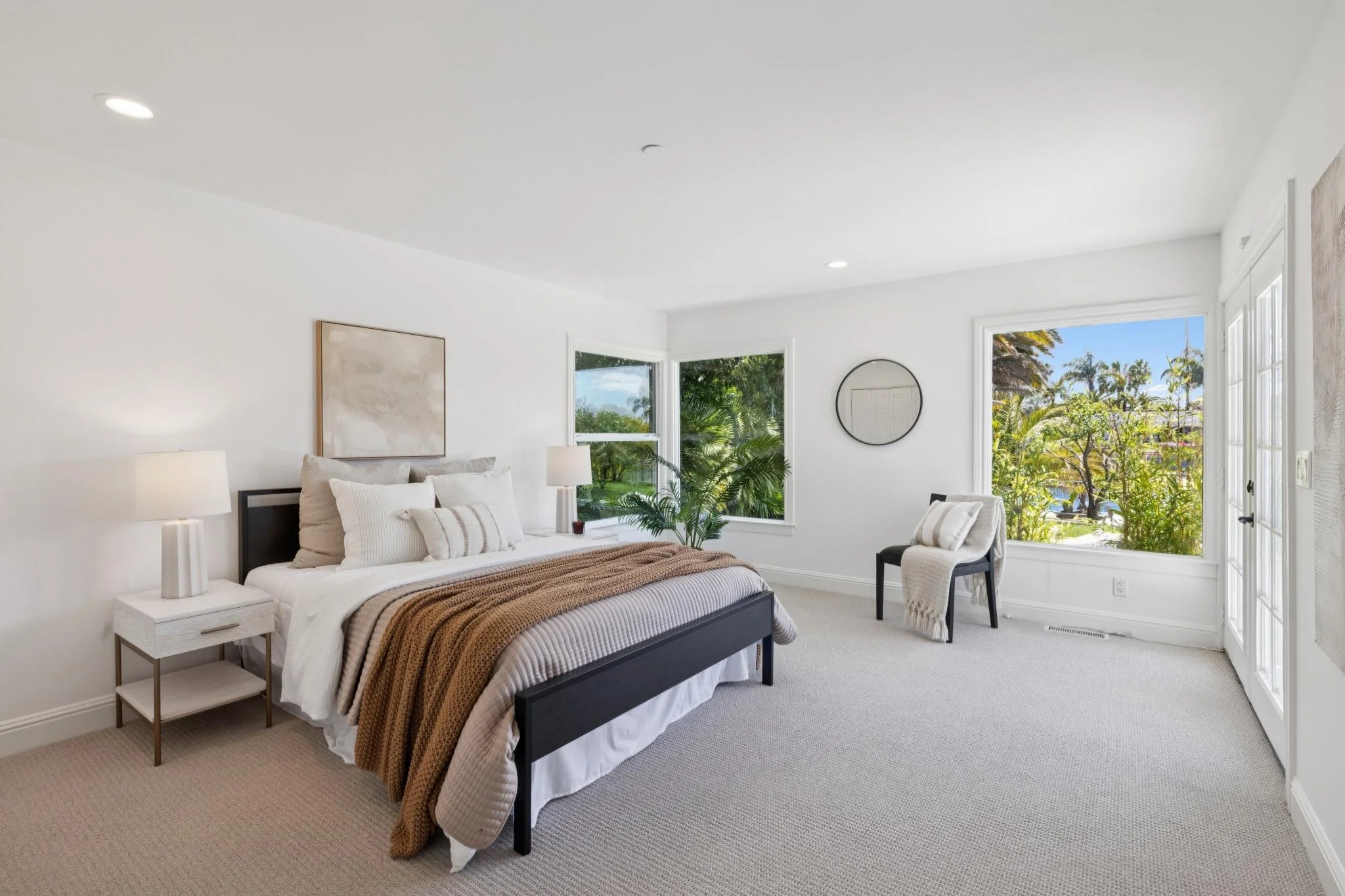 Bright bedroom with large windows showing greenery outside, a bed with white and brown bedding, nightstand with a lamp, and a sitting chair with a blanket.