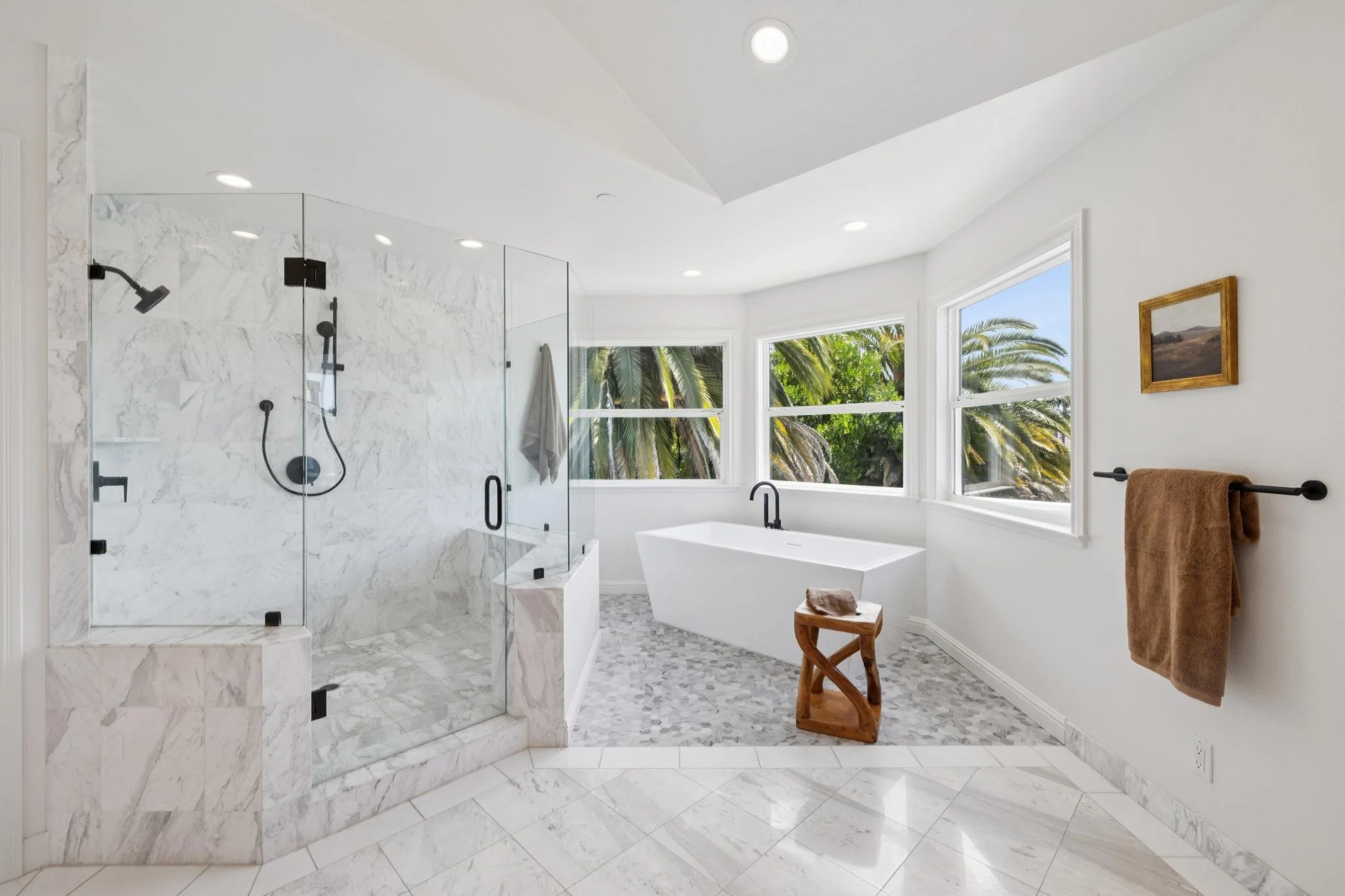 A modern bathroom with a marble shower and white bathtub near large windows with trees outside, a framed picture on the wall, and a brown towel on a towel rack.