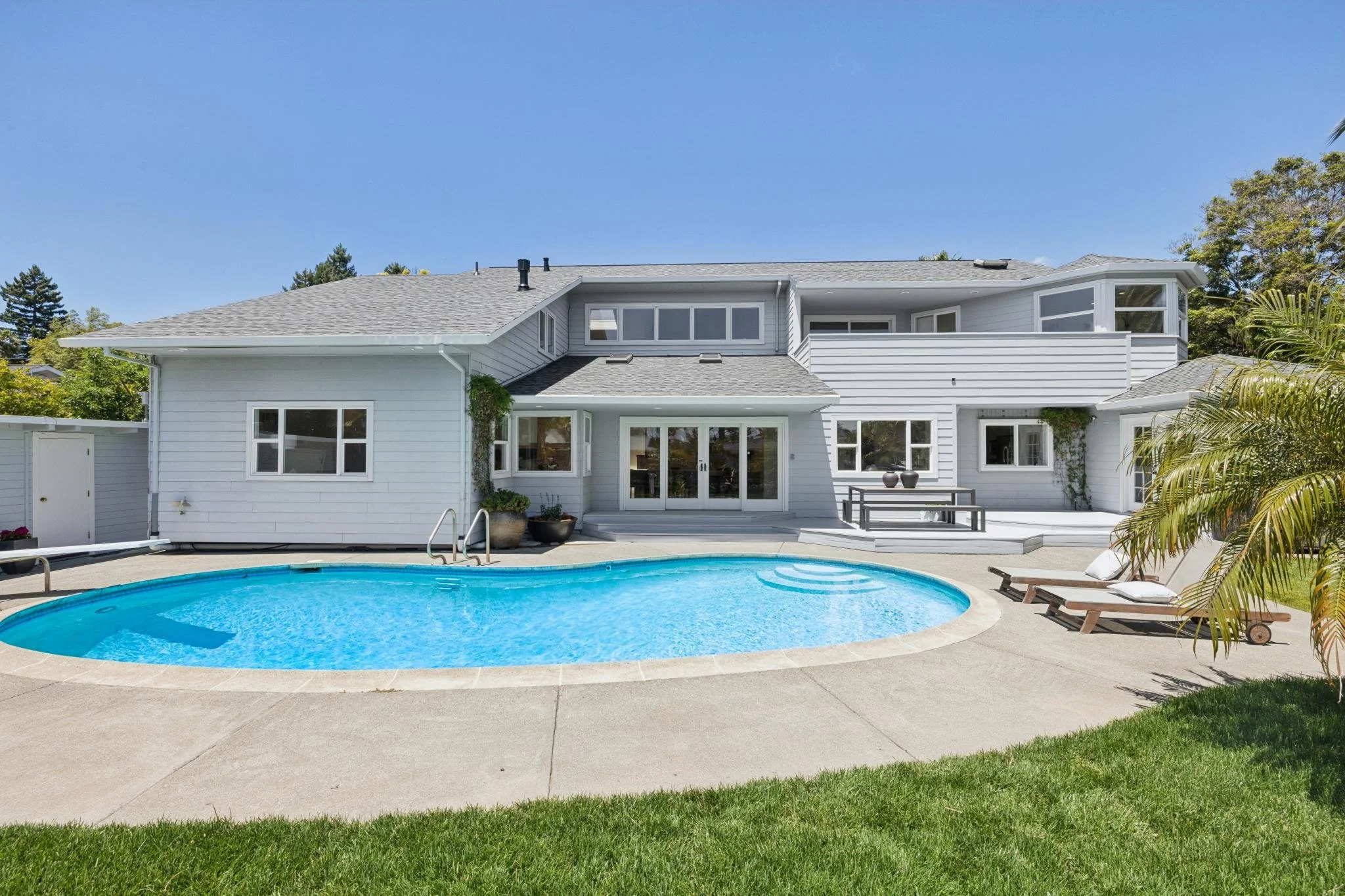A modern white two-story house with large windows, a backyard with a swimming pool, and outdoor seating areas. The house has a gray shingle roof and surrounding trees and plants under a clear blue sky.