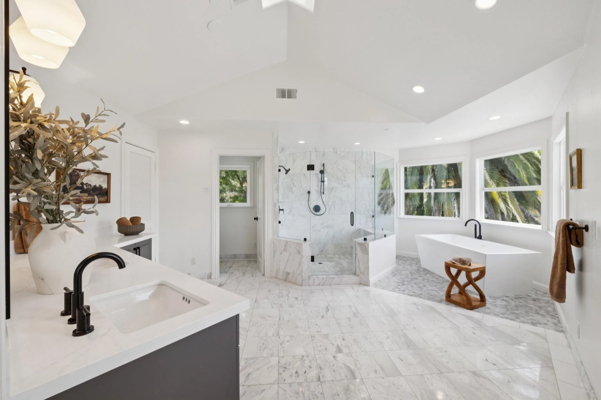 Bright modern bathroom with white marble floors and walls, large windows with greenery outside, a glass shower enclosure, a bathtub, and black fixtures.