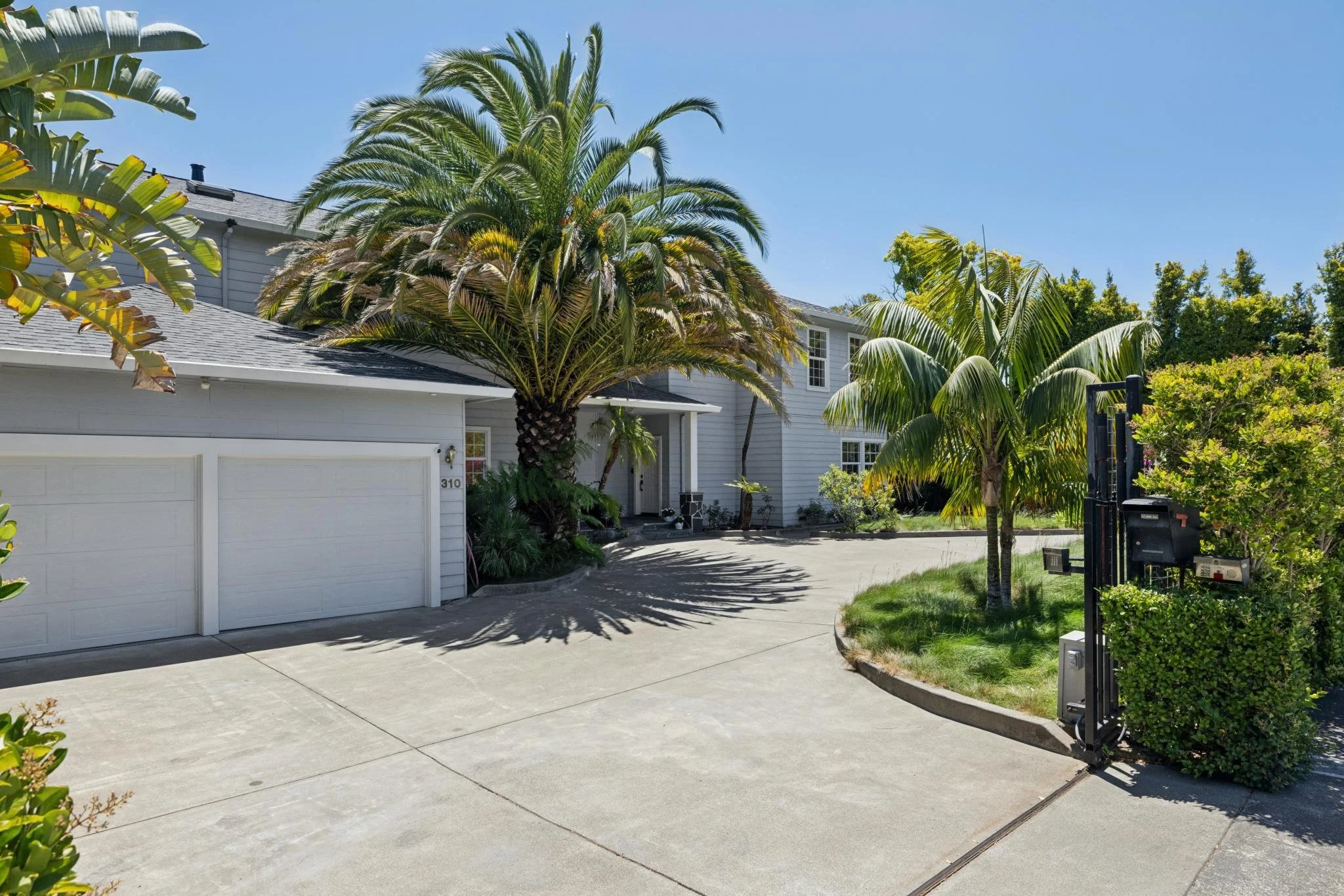 A house with a gray exterior and a double garage door surrounded by tropical trees and plants, including a large palm tree, with a clear blue sky above.