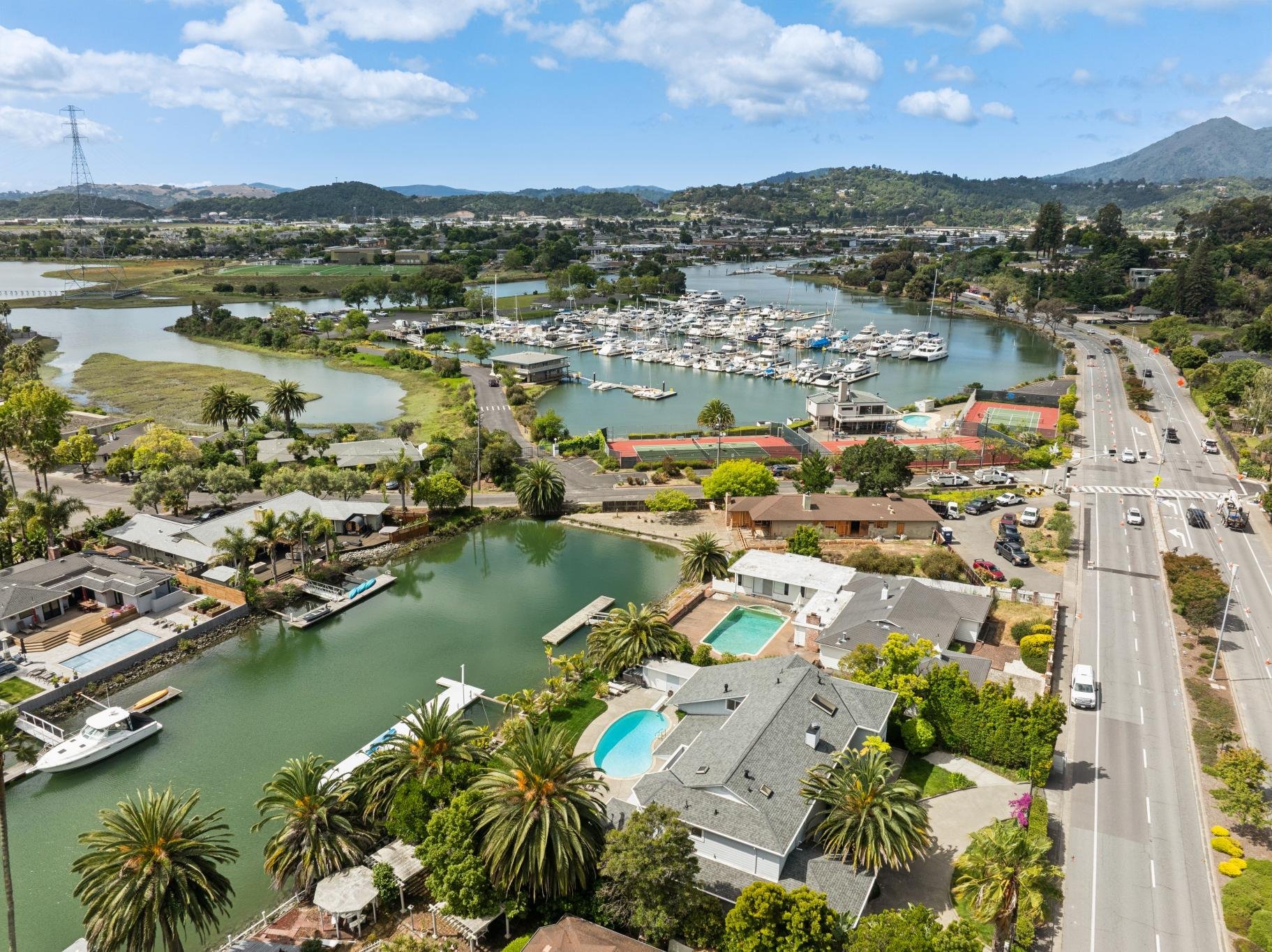 Aerial view of a marina with boats, houses with backyards, and a road lined with trees on a sunny day with blue skies and some clouds.