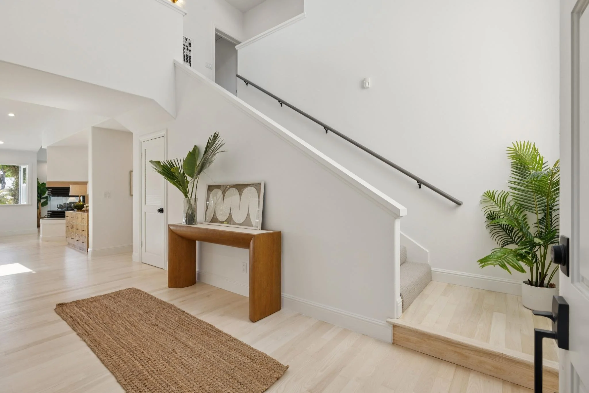 Modern interior home foyer with wooden console table, potted plants, white walls, staircase, and natural light.