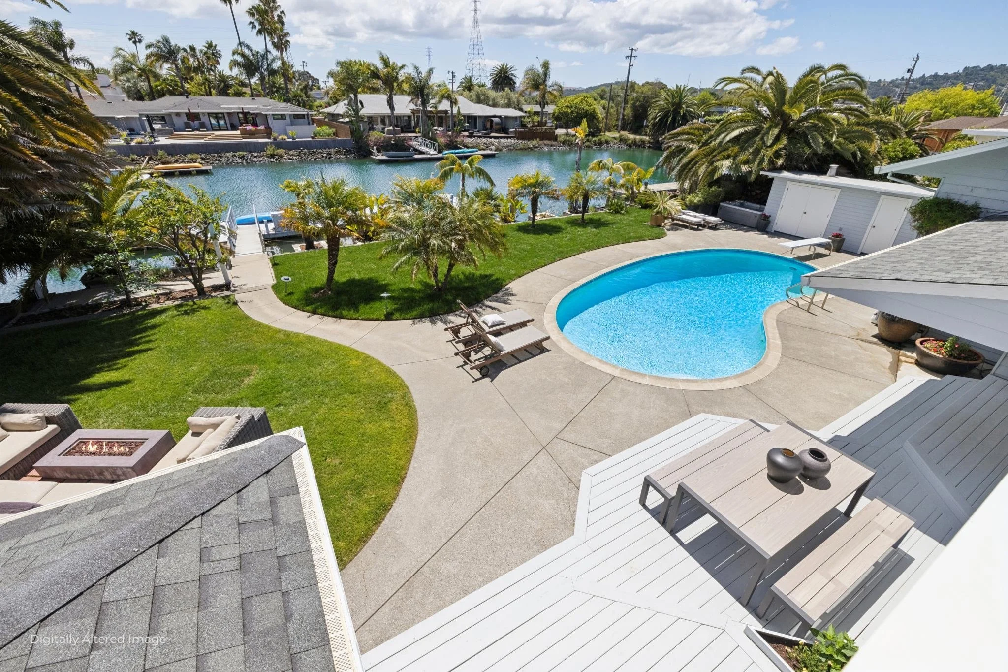 A backyard with a swimming pool surrounded by lounge chairs, a grassy area with small palm trees, and a view of a canal with houses and boats on the opposite side.