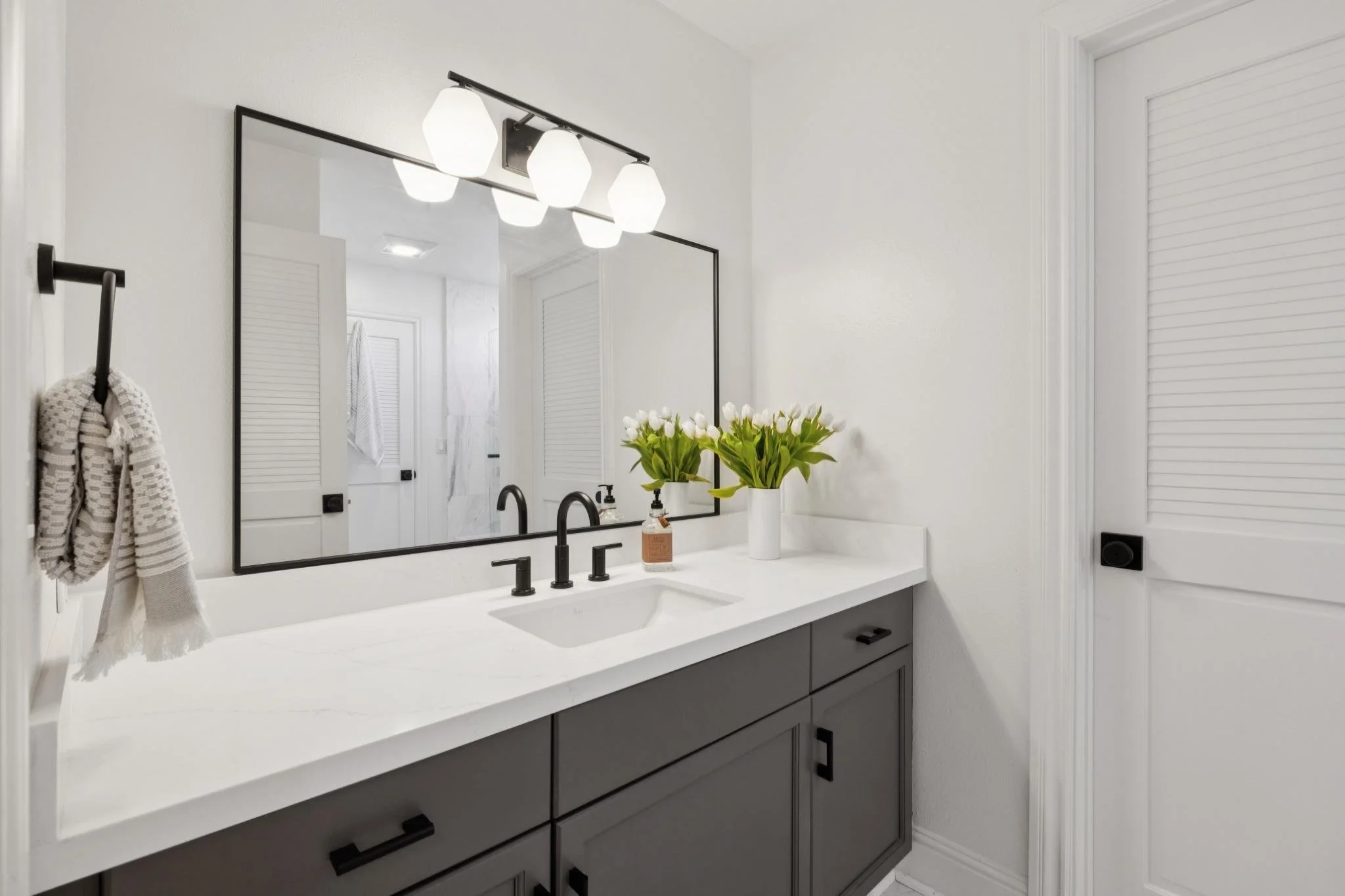 Modern bathroom with white walls and countertop, black fixtures, large mirror, LED light fixture, and a vase with white tulips on the counter.