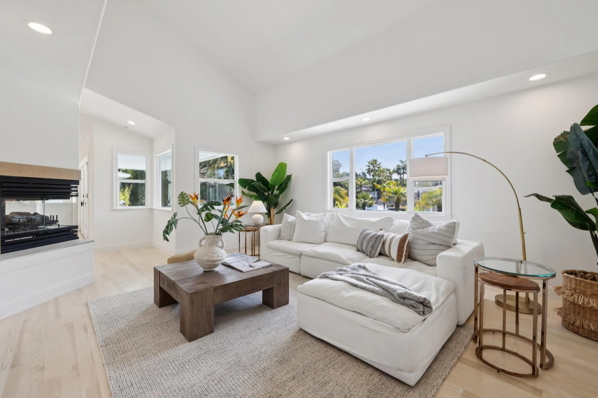 Bright living room with white walls, large windows showing outdoor greenery, a white sectional sofa with mixed pillows, wooden coffee table with a large vase, indoor plants, and a floor lamp.