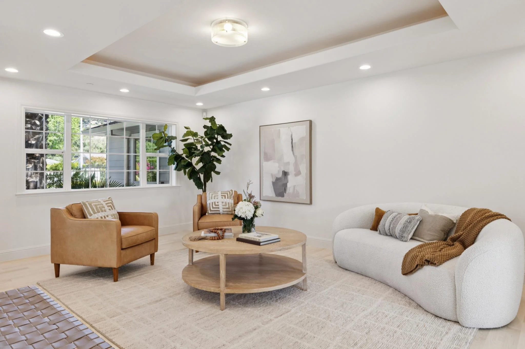 Living room with white walls, large window, brown leather armchairs, a white curved sofa with pillows, a round wooden coffee table with books and decorations, a large plant, and a piece of abstract artwork on the wall.