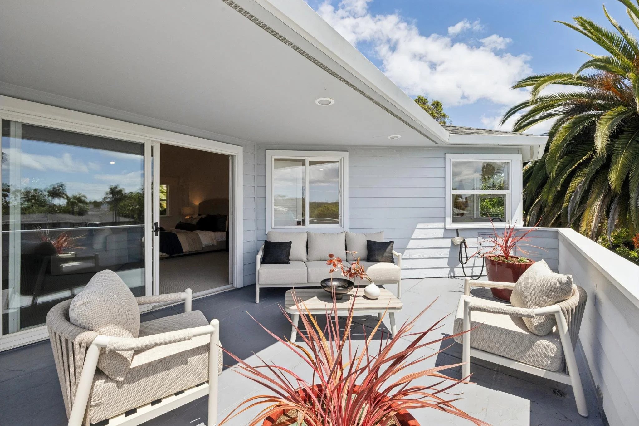 View of a balcony with outdoor furniture, a large potted plant, and a sliding glass door leading inside a bedroom with a bed and bedside lamp. The balcony has a white railing and modern decor, and is adjacent to a house with light blue siding, surrou