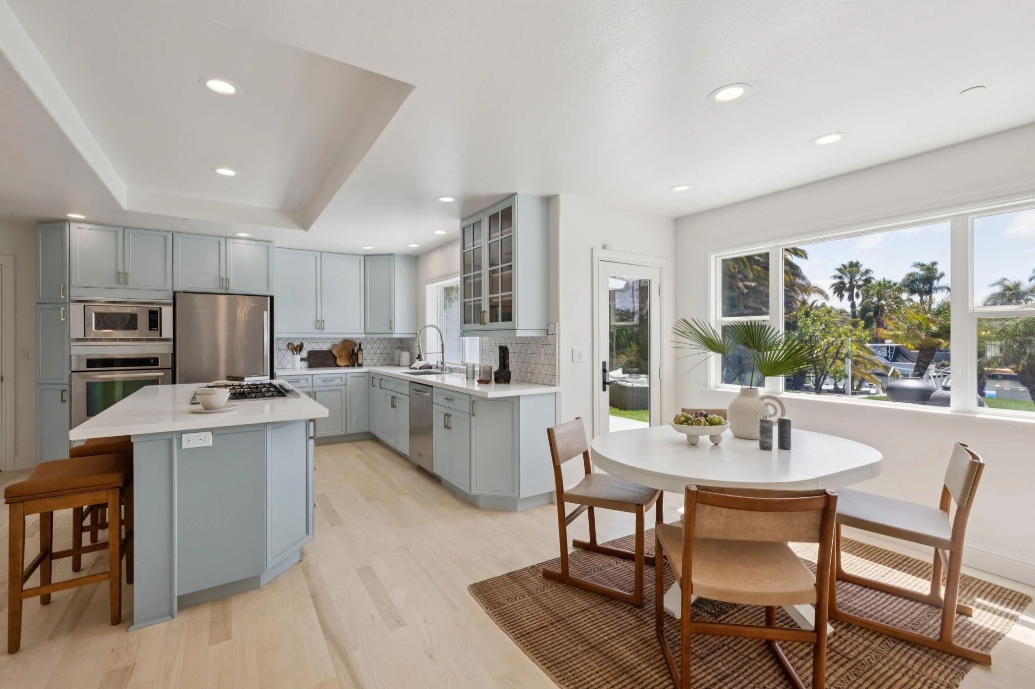 Bright kitchen with light blue cabinets, white countertops, and large windows overlooking a yard with palm trees. There is a round dining table with wooden chairs, and decorations including a white vase and a fruit bowl.
