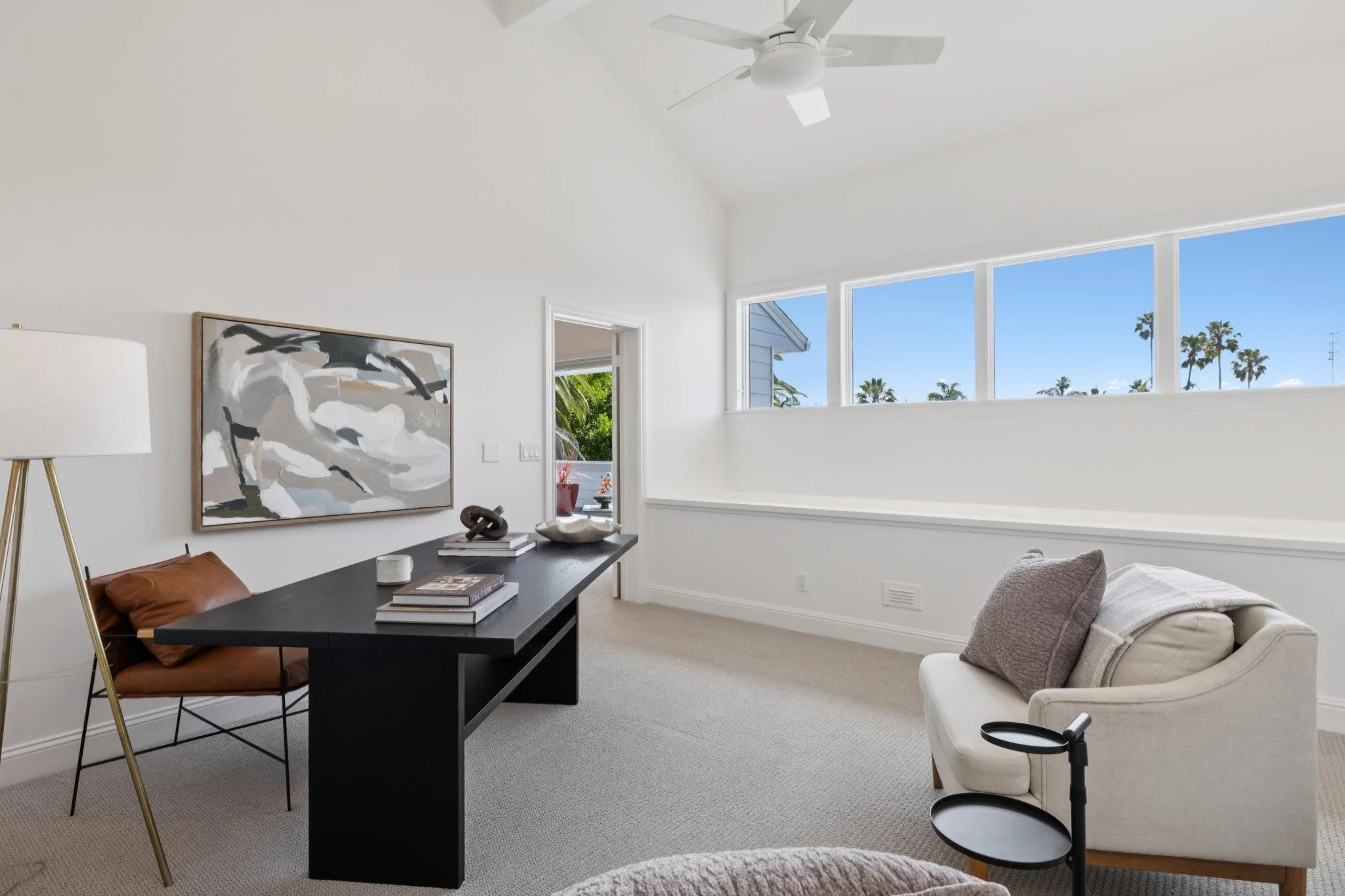 Living room with a white ceiling fan, a large window with a view of palm trees, a dark wood desk with books and decor, a tan leather chair, and a white armchair with pillows.