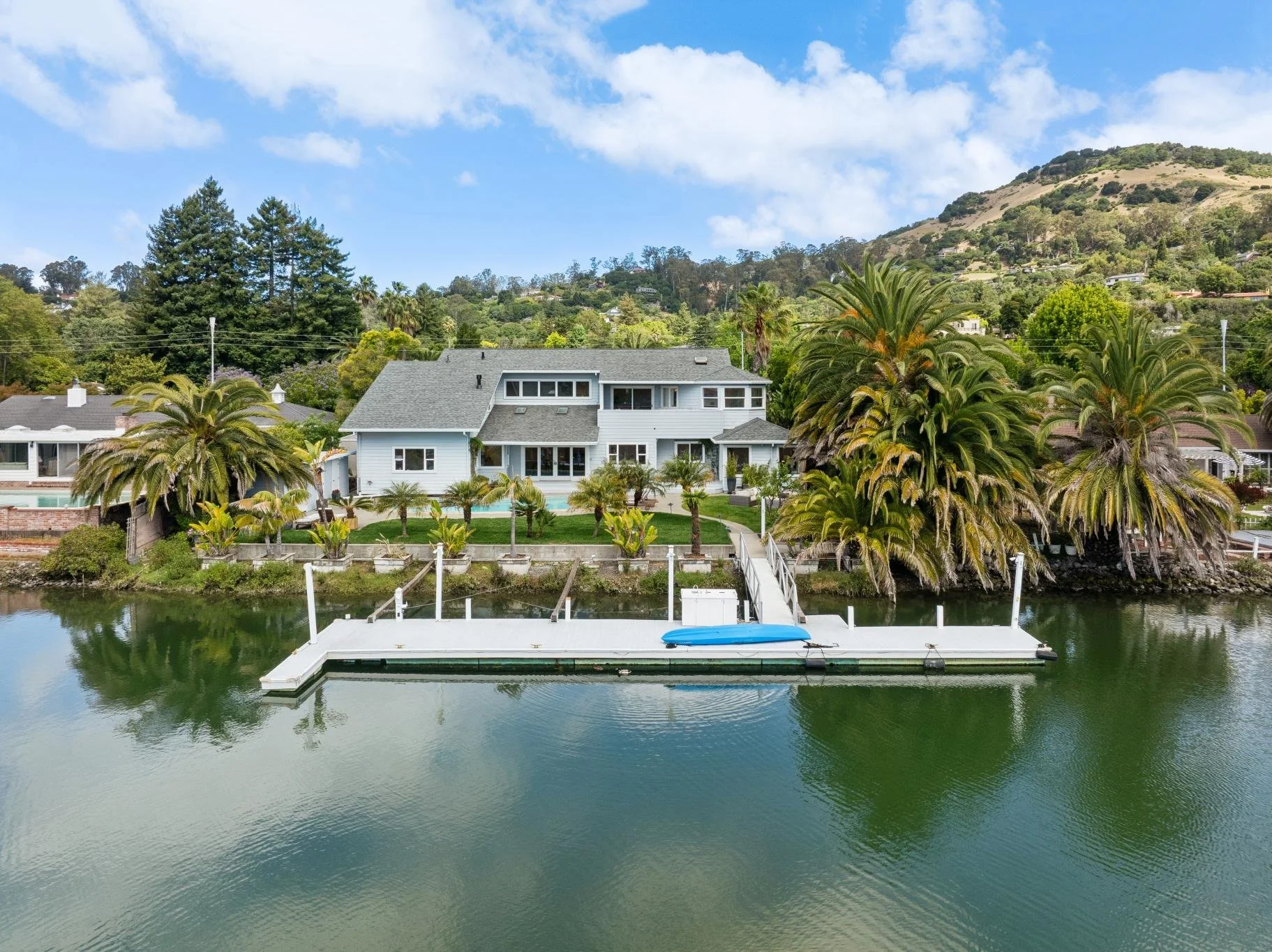 A house with a gray roof and white walls located near a waterway with a dock and a small boat, surrounded by lush palm trees and greenery, with a hill covered in trees in the background under a partly cloudy sky.