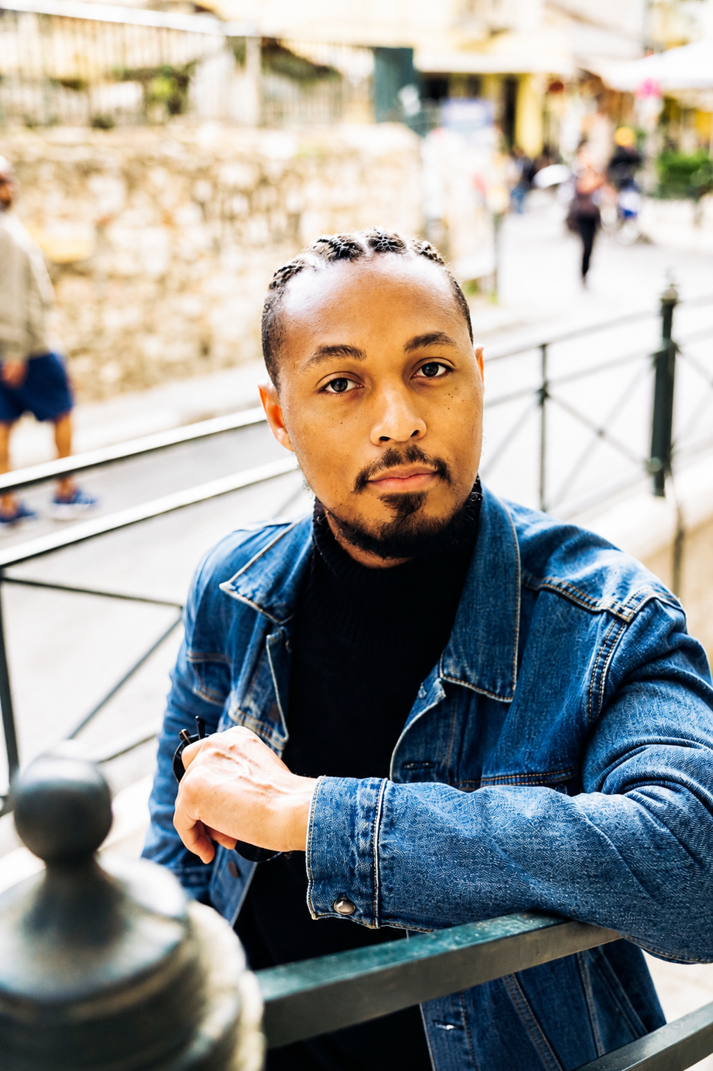 A man with brown skin, black hair styled in braids, and facial hair, wearing a blue denim jacket over a black shirt, sitting outdoors near a metal railing with a blurred urban background.