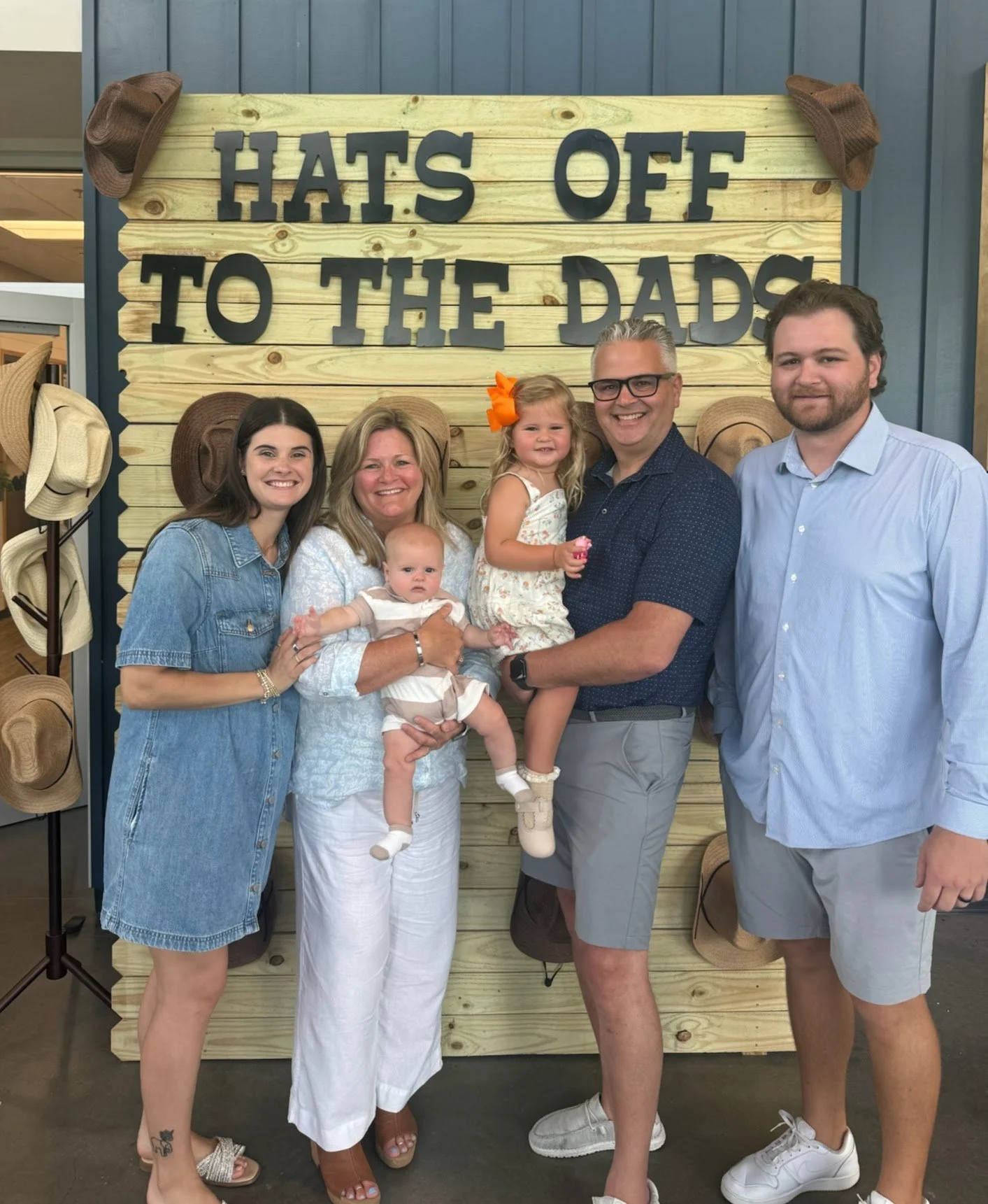 A group of six people, including two children, standing in front of a wooden sign that reads 'HATS OFF TO THE DADS'. The sign has hats hanging on its edges. The group is smiling and posing for a photo.