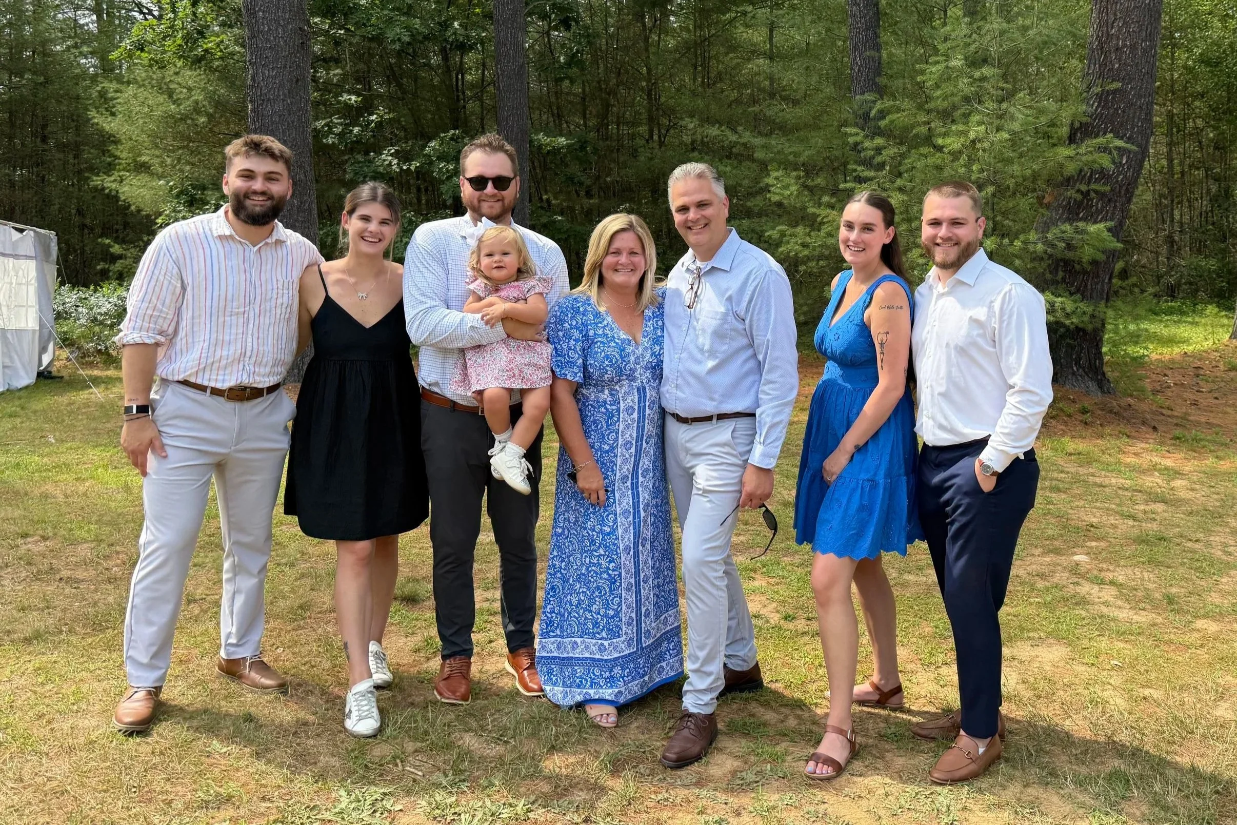 Group of nine people, including children, standing outdoors on grass with trees in the background, smiling for a photo on a sunny day.