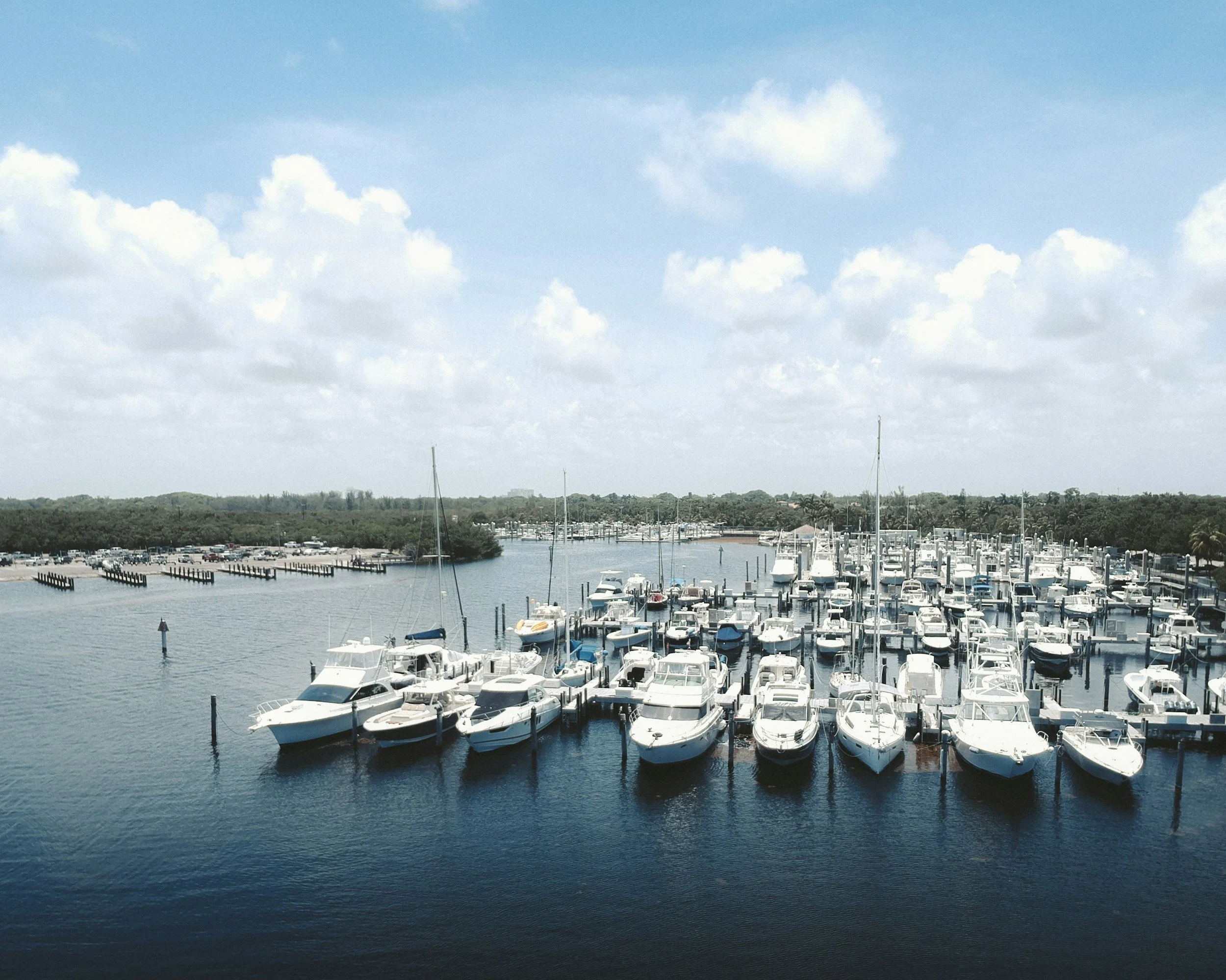 View of a marina with numerous boats docked, calm water, and a partly cloudy sky.