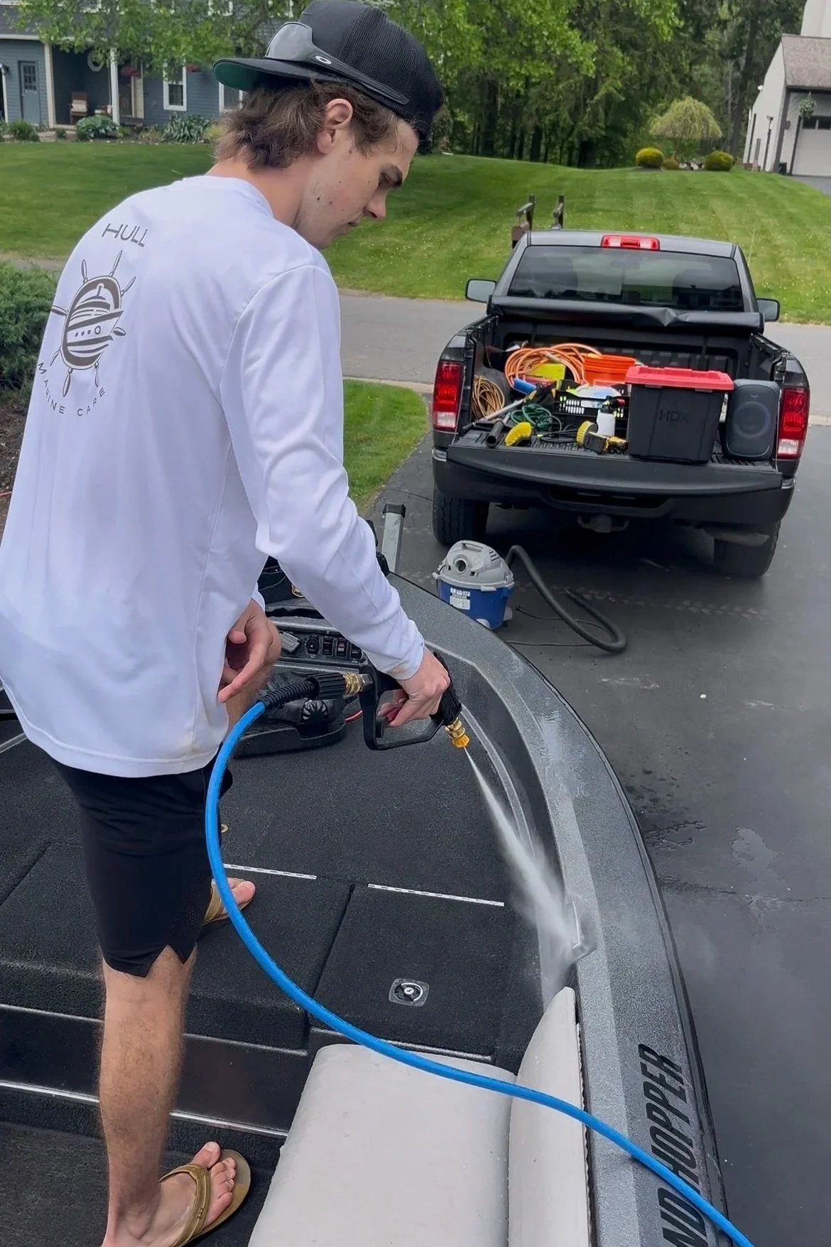 A young man is washing a boat with a pressure washer in a driveway. He is wearing a white long-sleeve shirt, black shorts, and sandals. The boat is on a trailer, and there is a black pickup truck in the background with tools and equipment in the truck bed.