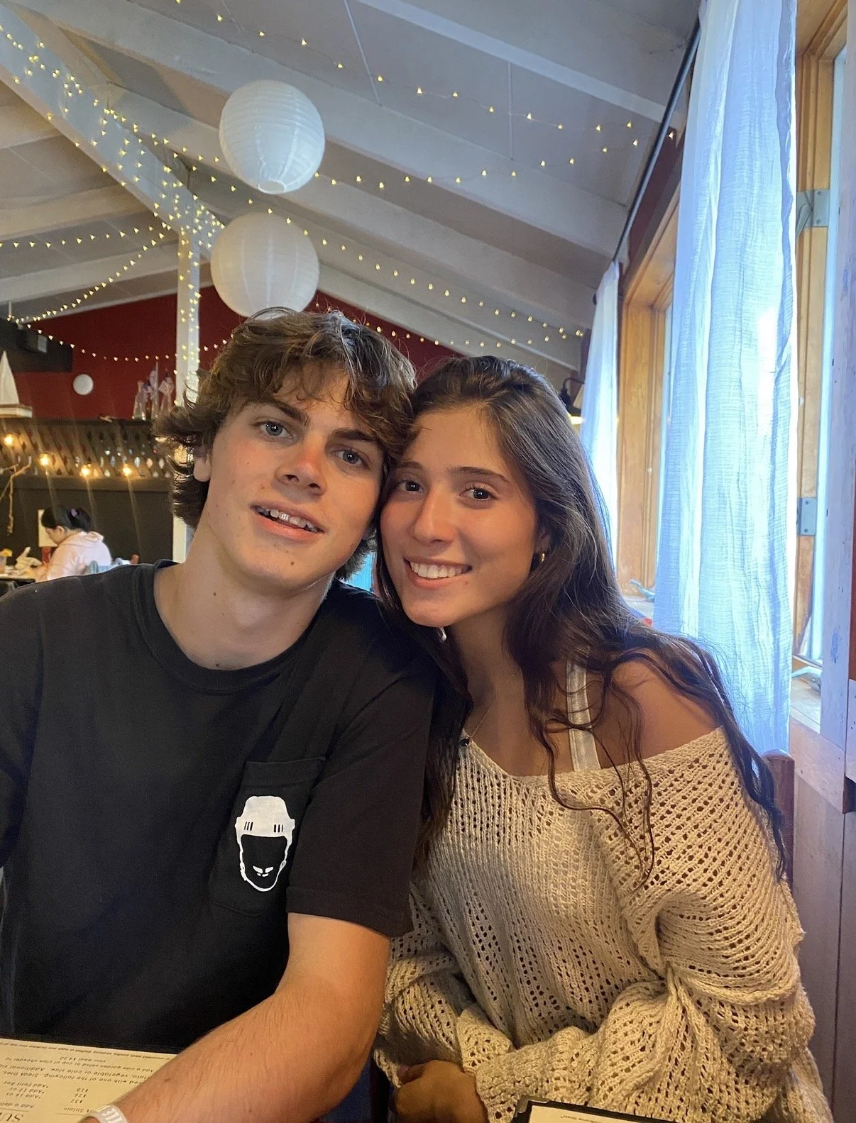 A young man and woman sitting closely together in a restaurant, smiling at the camera. The restaurant has hanging paper lanterns and string lights, with a window and curtains in the background.