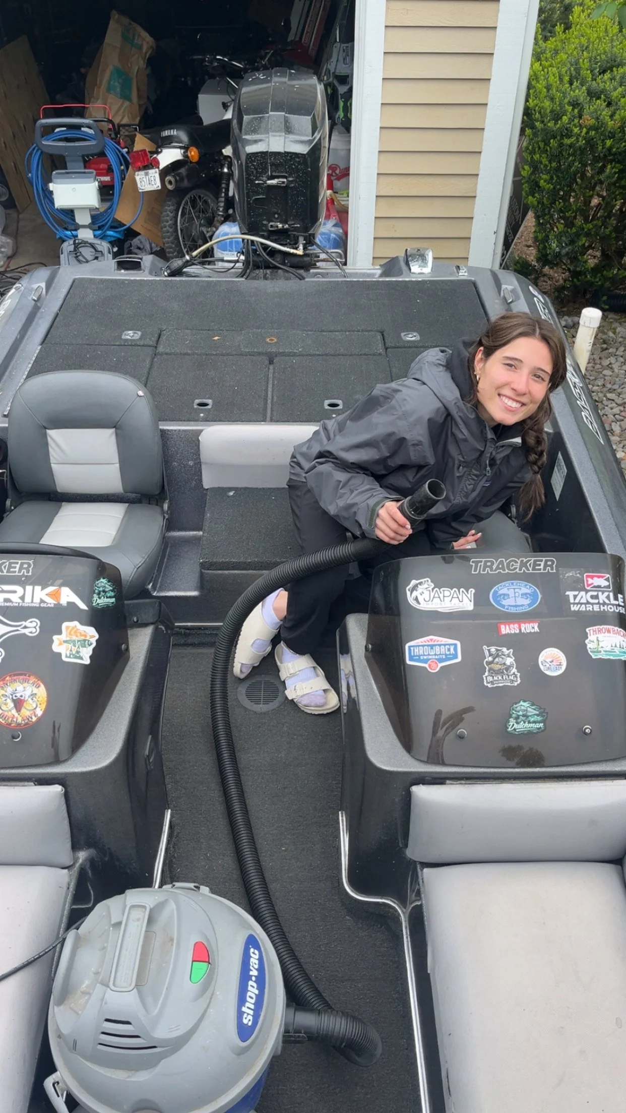 Smiling woman in a gray jacket sitting in a boat, holding a black piece, with a vacuum cleaner nearby, parked outside a garage with a motorcycle and various tools visible in the background.