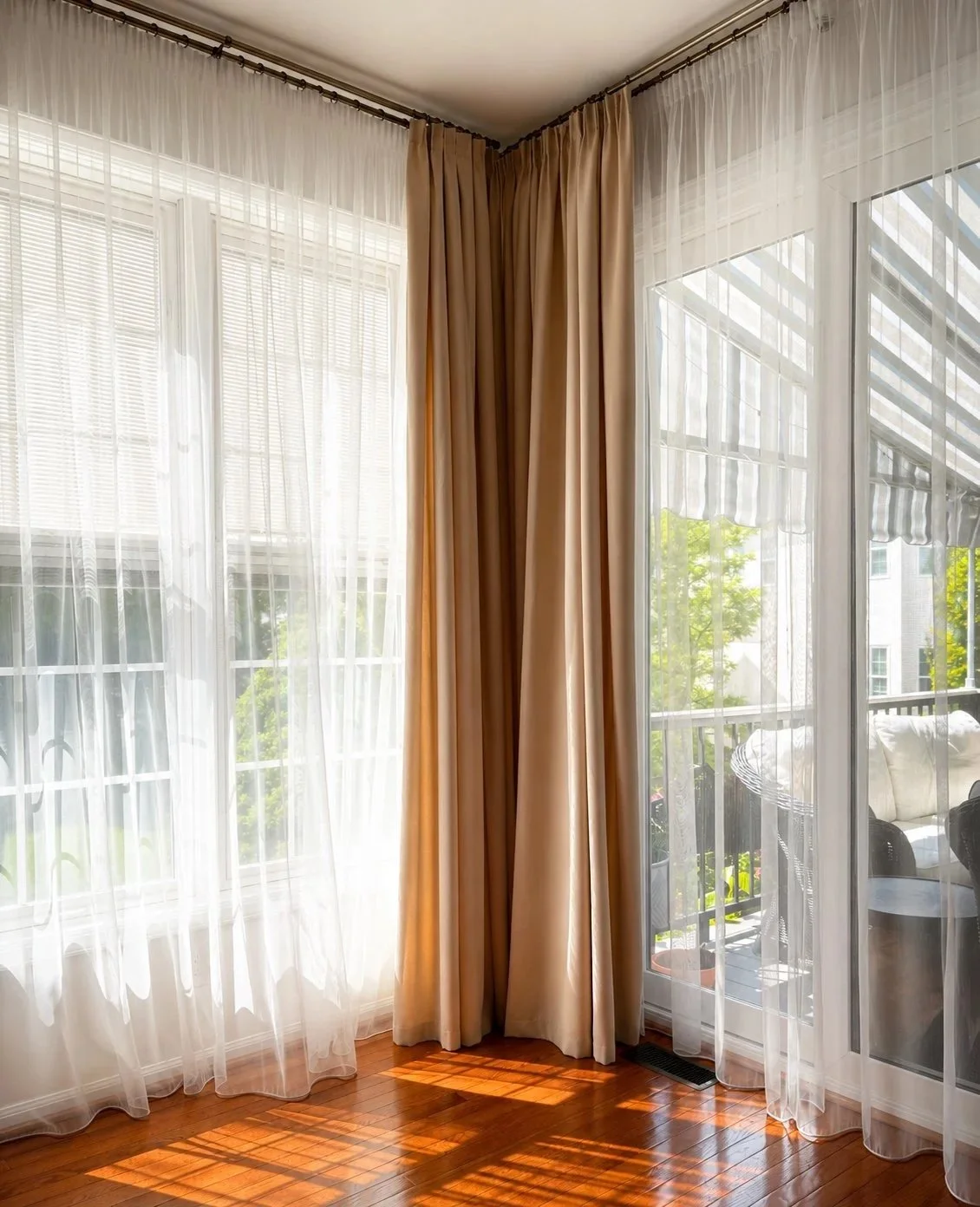 Sunlight streaming through sheer white curtains and beige drapes on a window and sliding glass door, with a balcony visible outside.