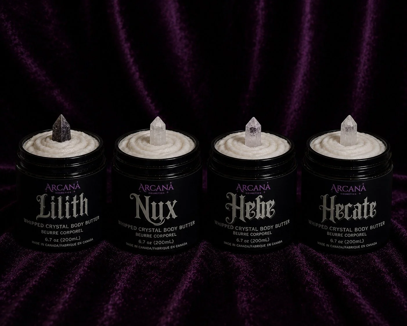 Four jars of Arcana Cosmetics whipped crystal body butter with crystal point toppers in black, clear, and frosted varieties, set against a dark purple velvet background.