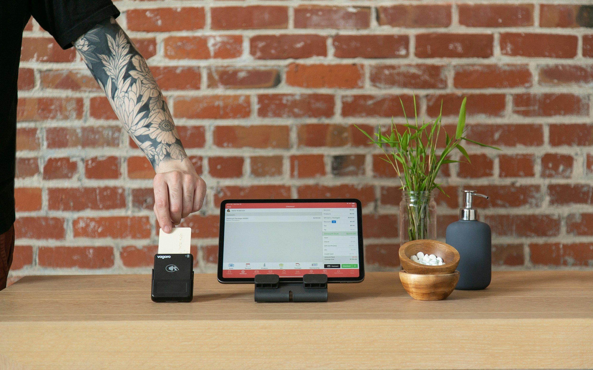 A person with a tattooed arm is checking out at a counter using a tablet device and a card payment terminal, with a brick wall in the background, a plant in a vase, a bowl of white pills, and a soap dispenser on the table.