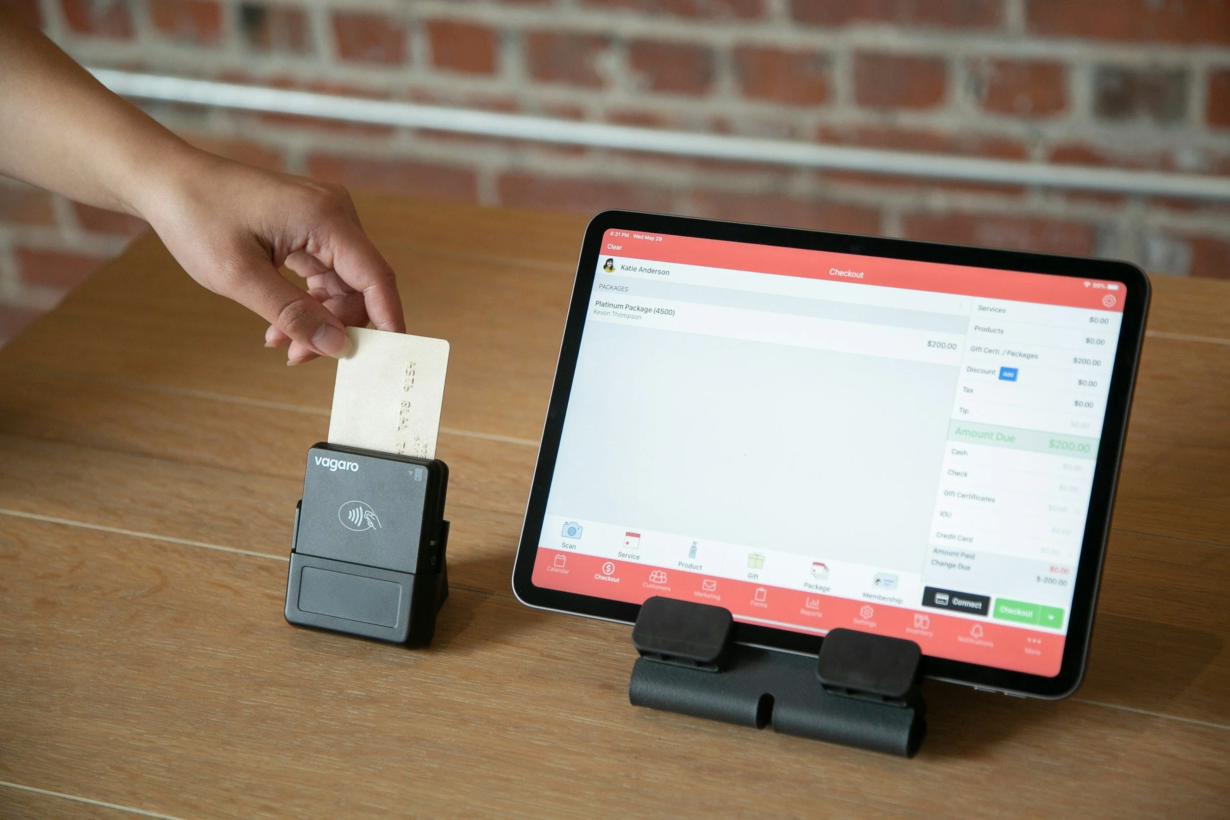A person is holding a gold-colored credit card near a card reader while a tablet screen displaying an online checkout process is on the table. The background has a brick wall.
