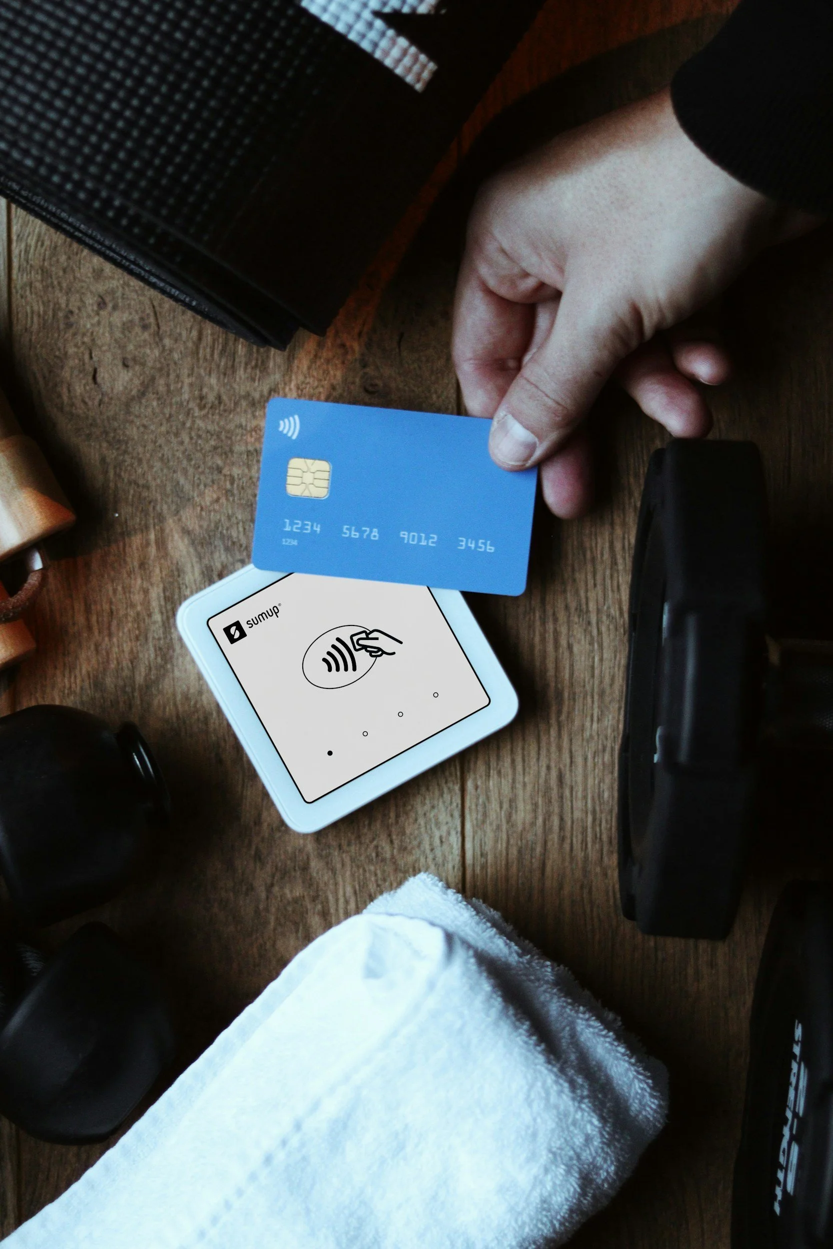 A person holding a blue contactless payment card over a white electronic payment device with instructions to tap. The scene is on a wooden surface with various objects including a black bag, a black camera, and a white towel.