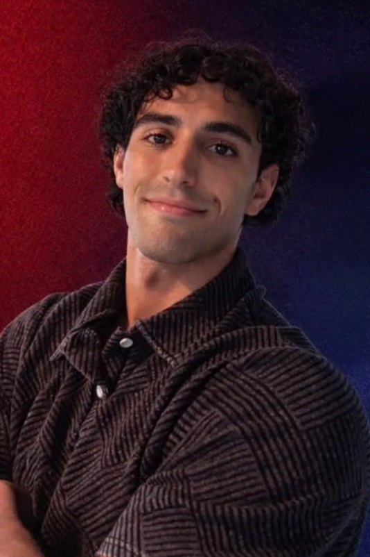 A young man with curly dark hair and a light smile, wearing a dark, patterned button-up shirt, posing in front of a red and blue background.