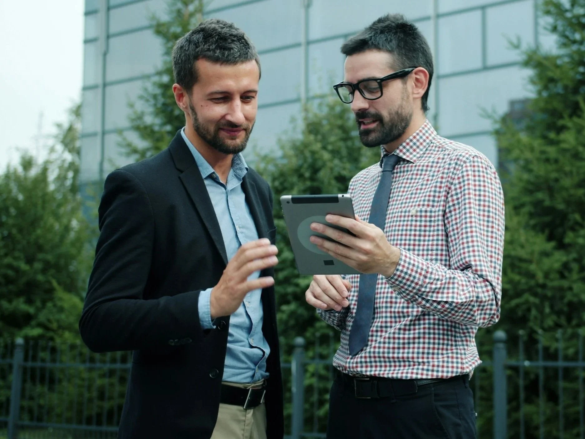 Two businessmen standing outdoors, looking at a tablet together. One is dressed in a black suit with a blue shirt, and the other wears glasses, a checkered shirt, and a tie. They are engaged in conversation.