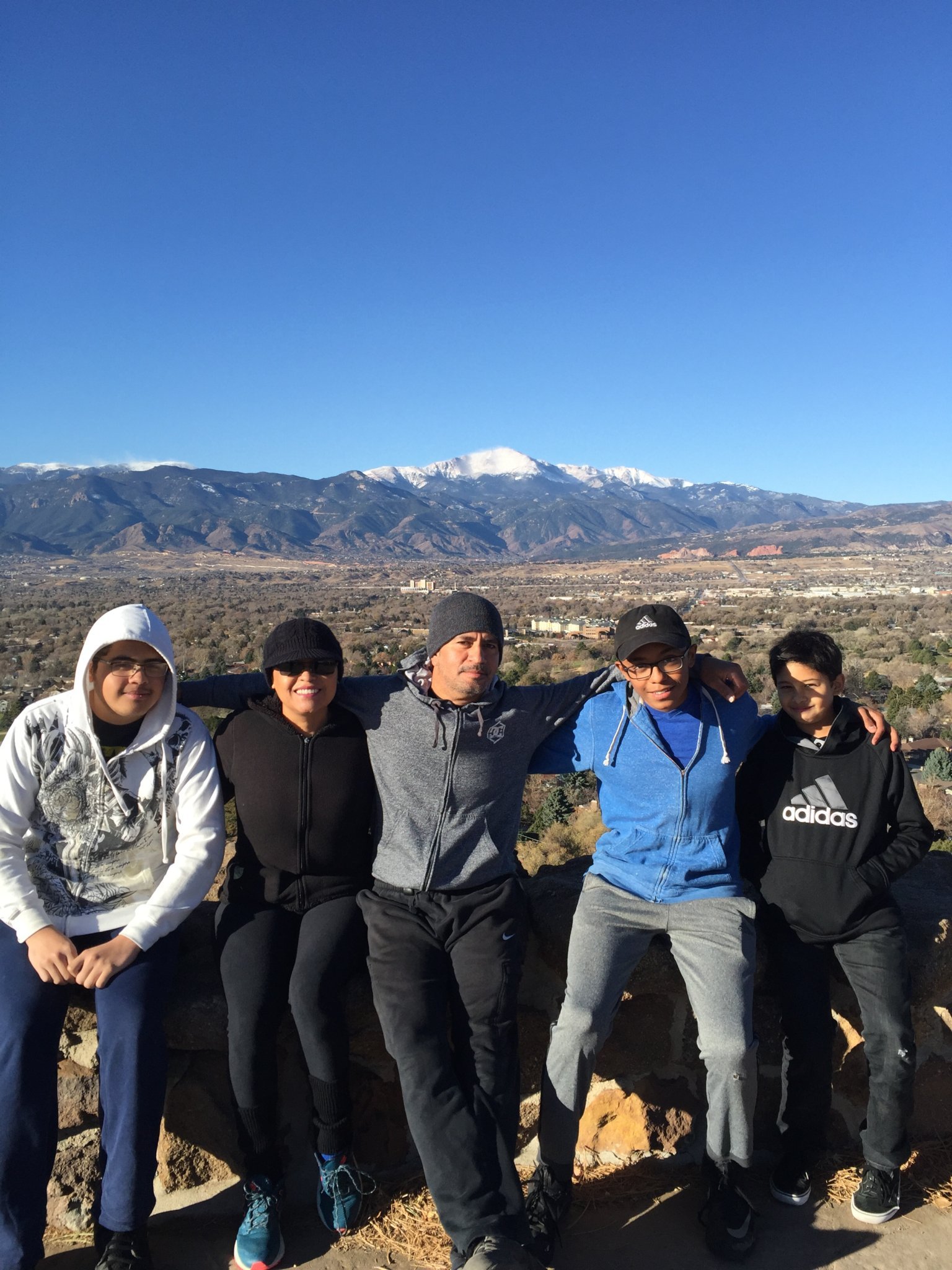 Group of five people standing outdoors in front of mountains with snow, clear blue sky, and a valley below, posing for a photo.
