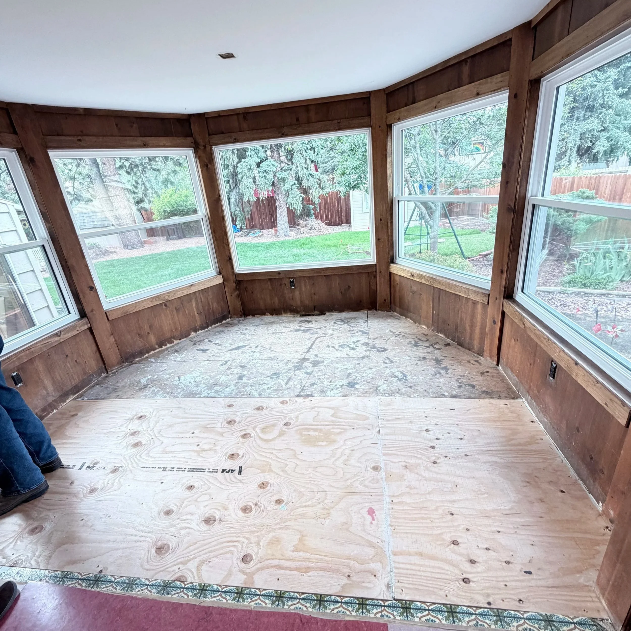 Interior of a room under construction with large windows, wood framing, and a plywood subfloor, outside view shows trees and a fenced backyard.