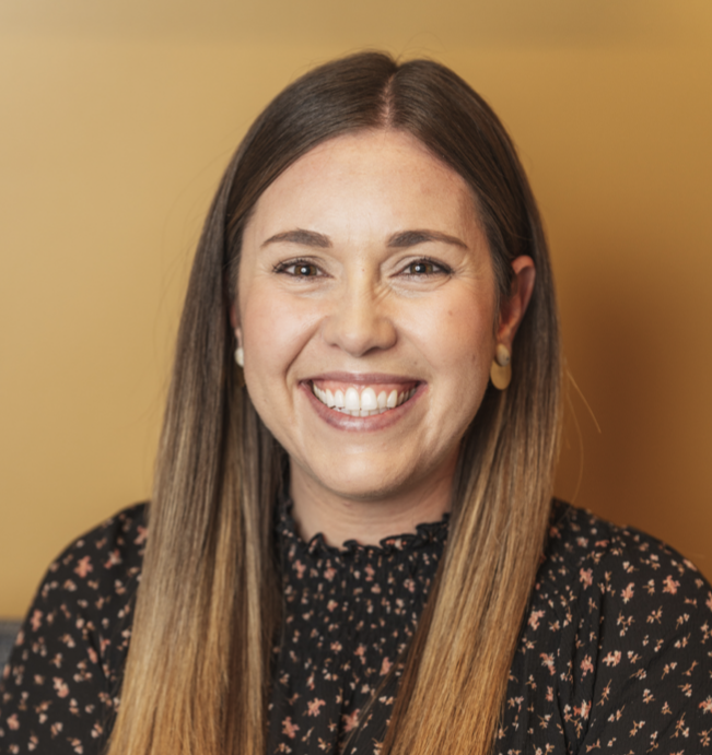 A woman with long brown hair smiling against a yellow background, wearing a black floral blouse and gold earrings.
