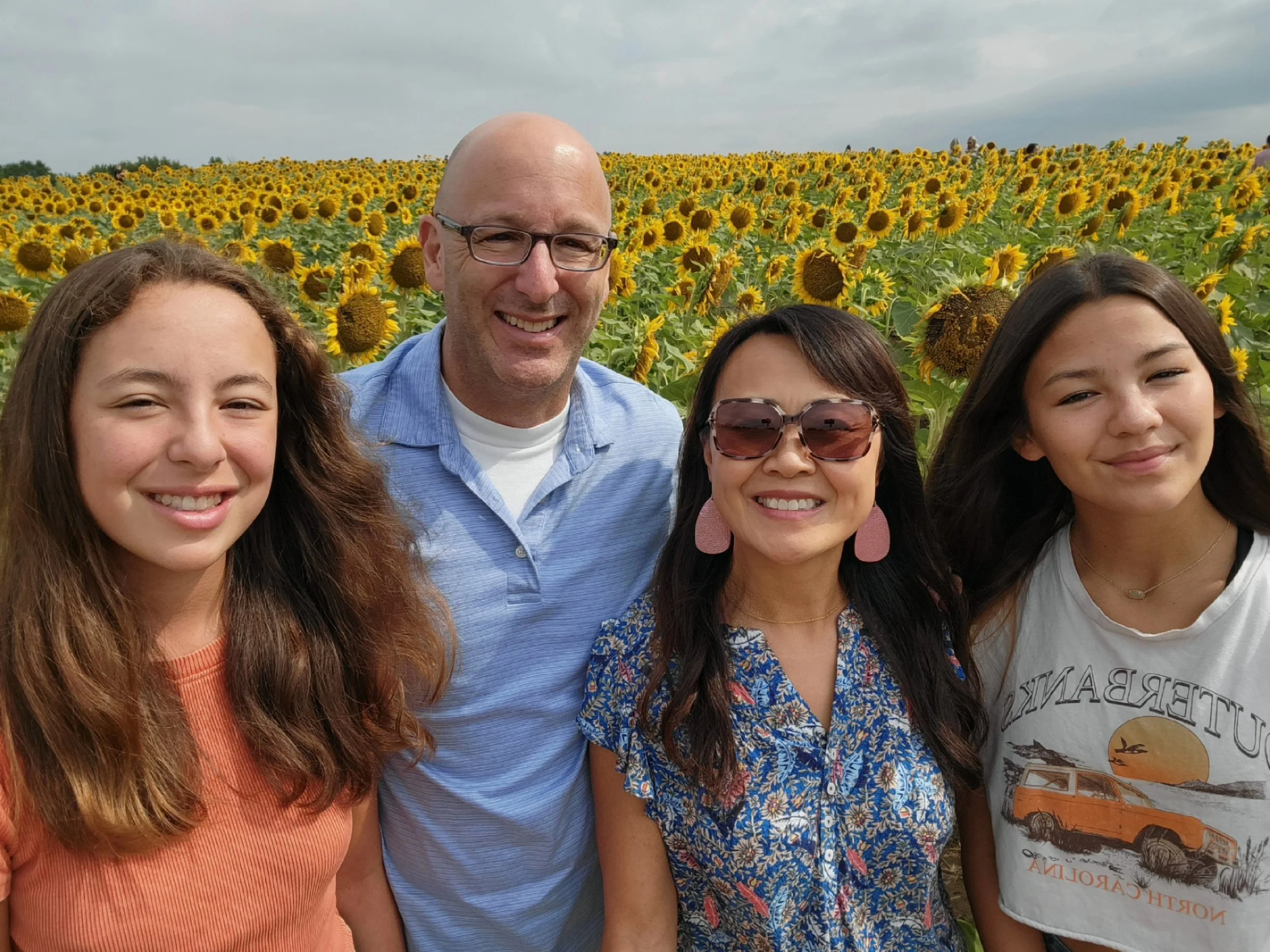 A group of four people smiling and taking a selfie in a sunflower field on a cloudy day.