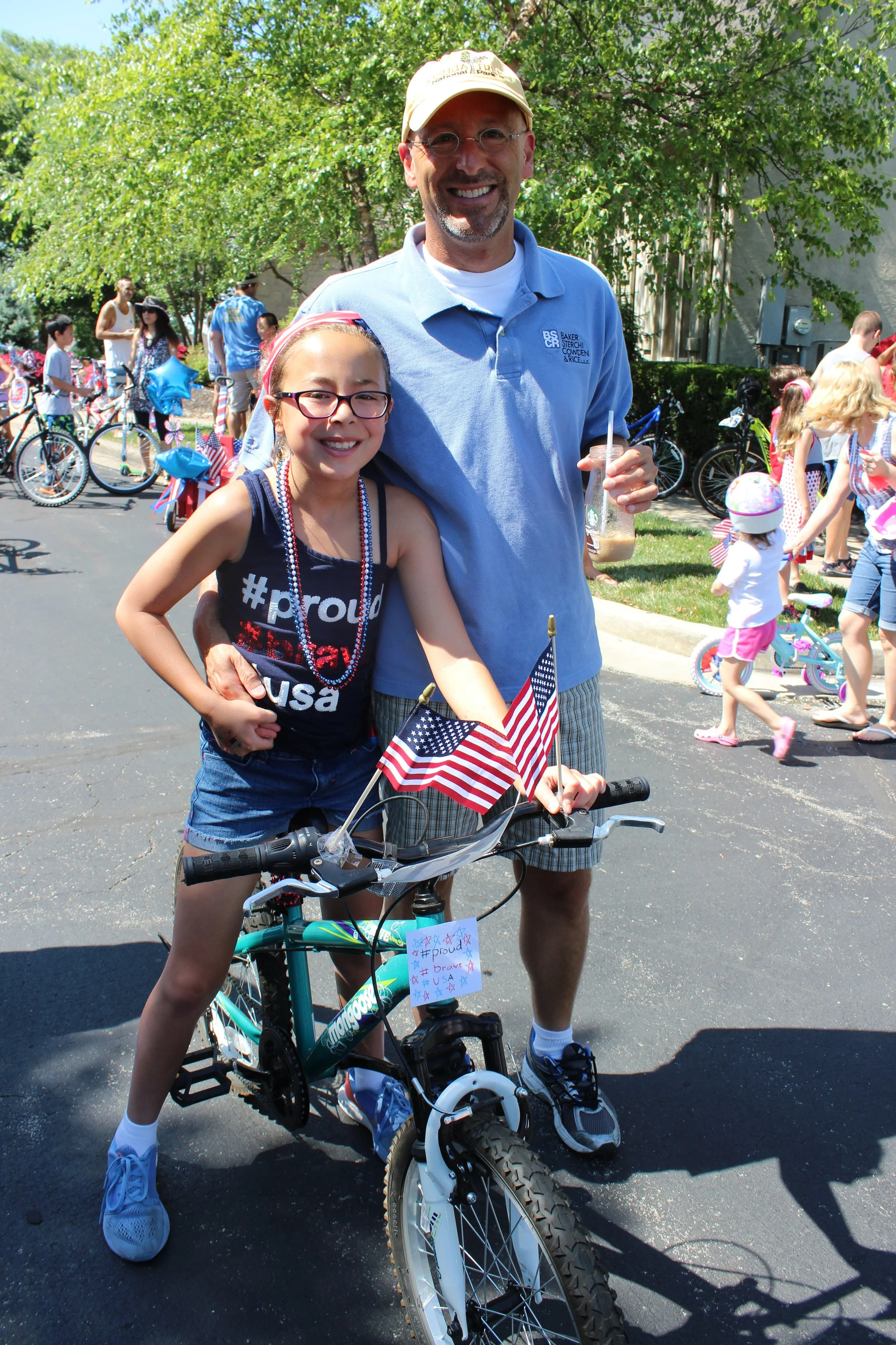 A man and young girl smiling at the camera indoors at a summer event. The girl is standing on a bicycle with an American flag attached, while the man stands beside her holding a drink. Children and bicycles are in the background.