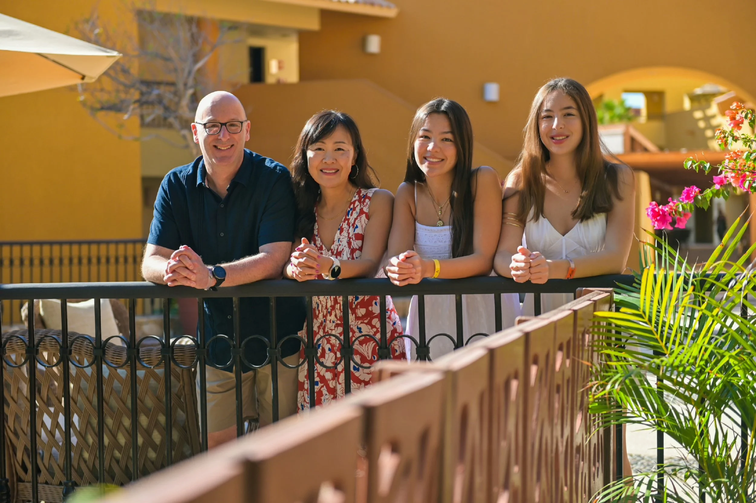 A group of four people, two women and two men, smiling and leaning on a balcony railing in an outdoor setting with yellow walls, a tree, and potted plants.