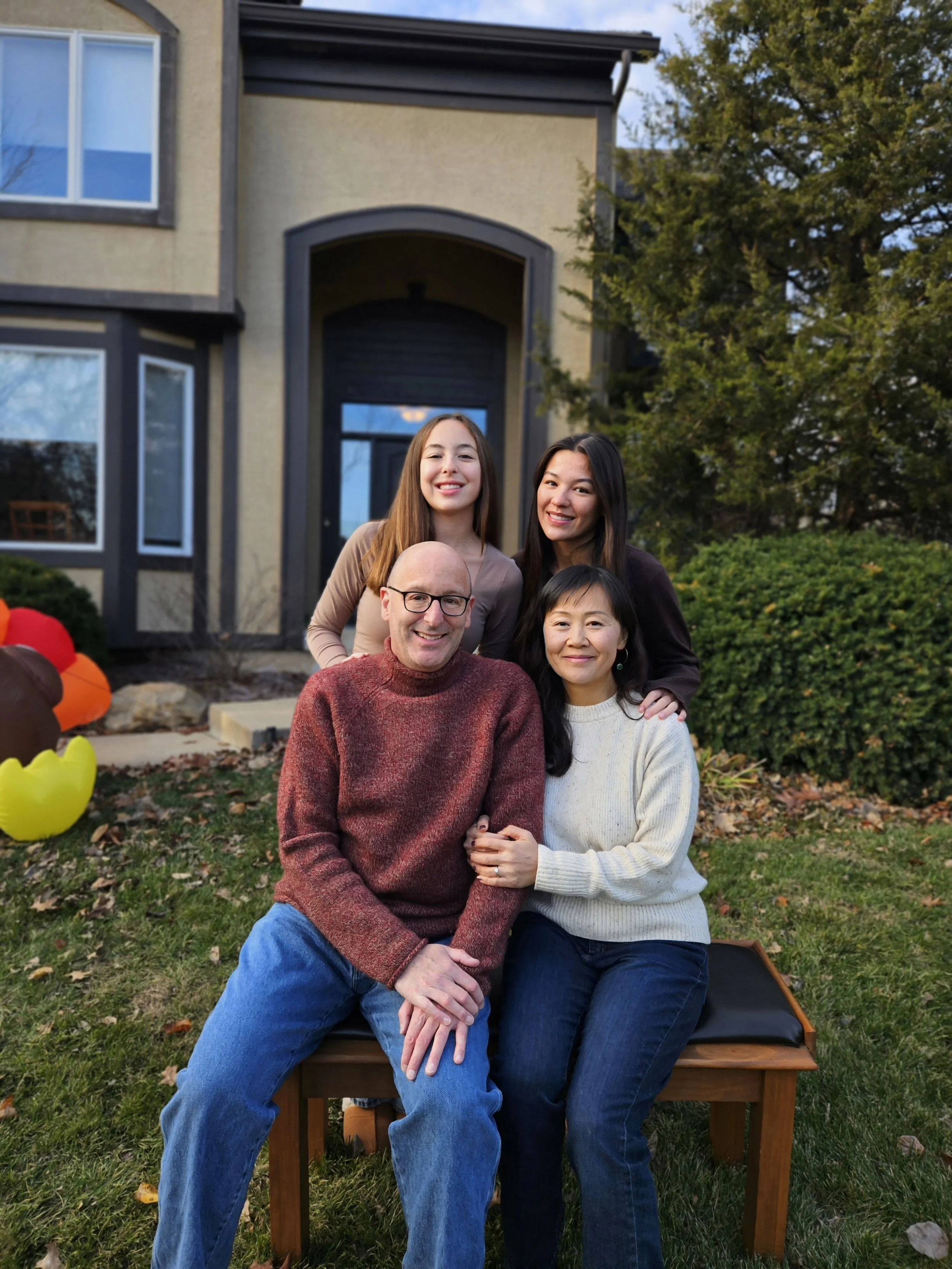 A group of five people, including two women, a man, and two children, sitting and standing on a wooden bench outside a house with large windows and a tree in the background. They are smiling and posing for the photo.