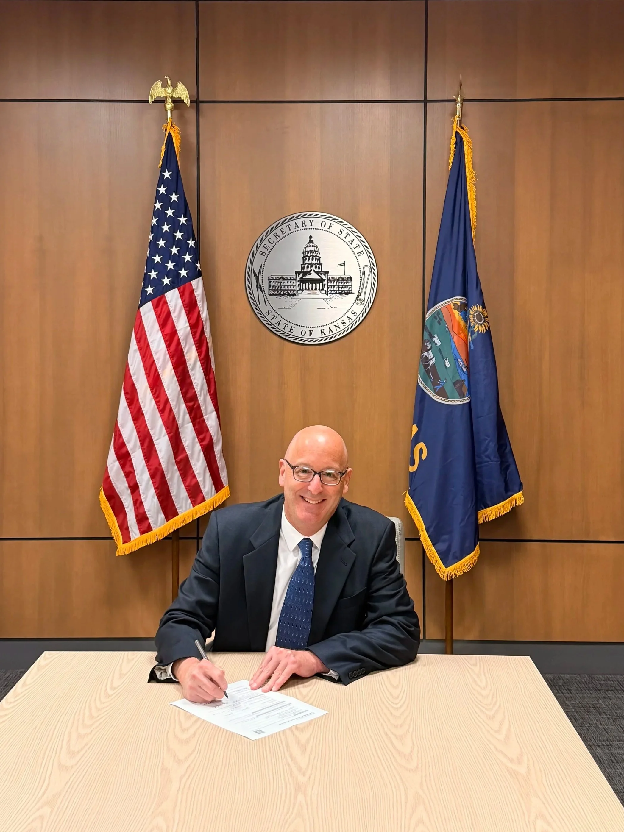 A man in a suit and tie sitting at a wooden table, signing a document with an American flag on his right, the flag of Kansas on his left, and the seal of the State of Kansas behind him. The wall behind him is made of wood panels.