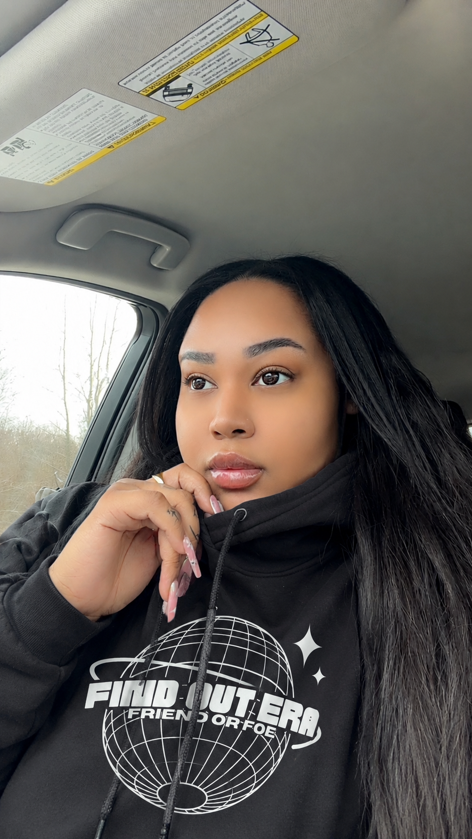 A young woman with long black hair sitting in a car, wearing a black hoodie with a graphic print that says "FIND OUT ERA FRIEND OR FOE" and looking out the window with a thoughtful expression.