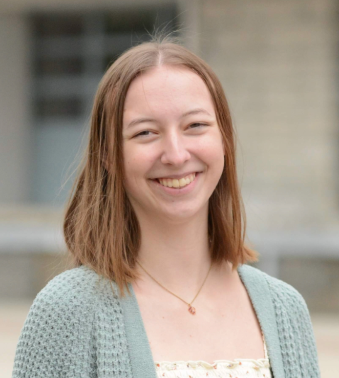 A young woman with shoulder-length light brown hair smiling outdoors, wearing a light green cardigan and a necklace with a small pendant.