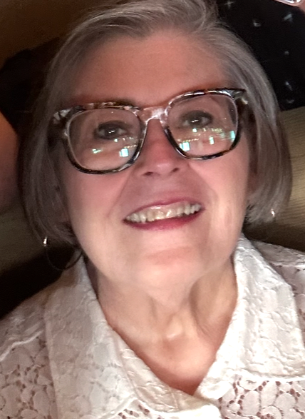 Close-up of an elderly woman with short gray hair and glasses, smiling and wearing a light-colored lace shirt.