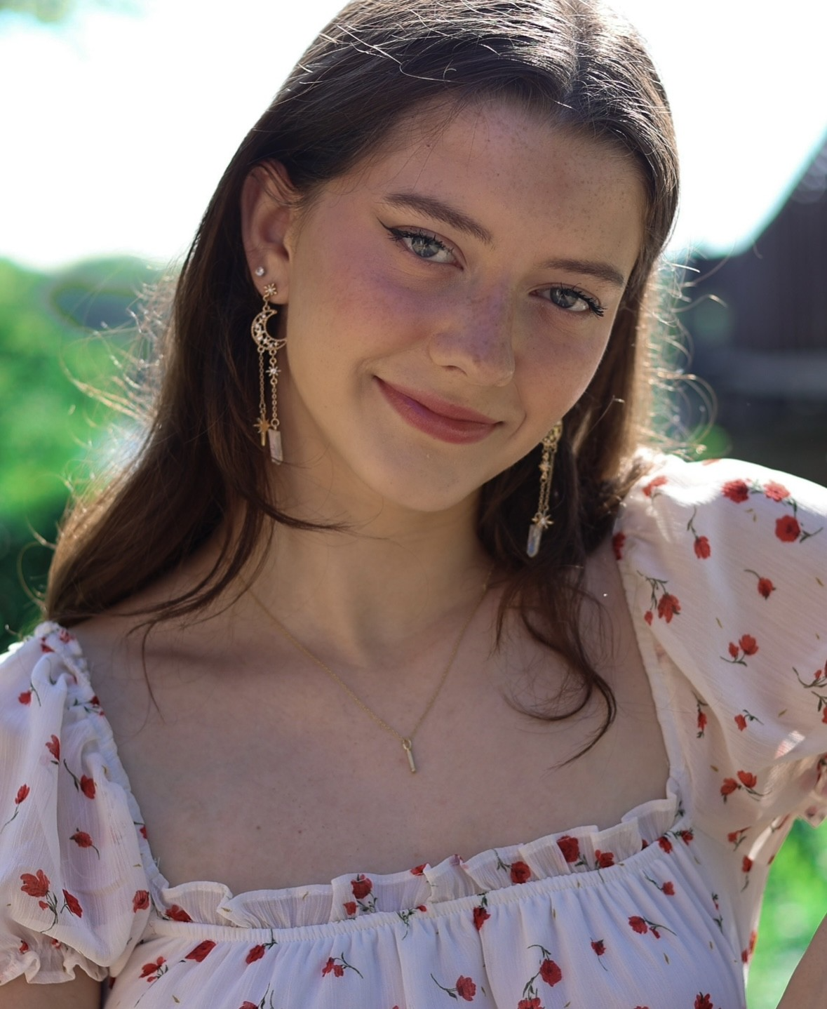 A young woman with long brown hair, wearing a white dress with red floral prints and gold jewelry, smiling outdoors with sunlight in the background.