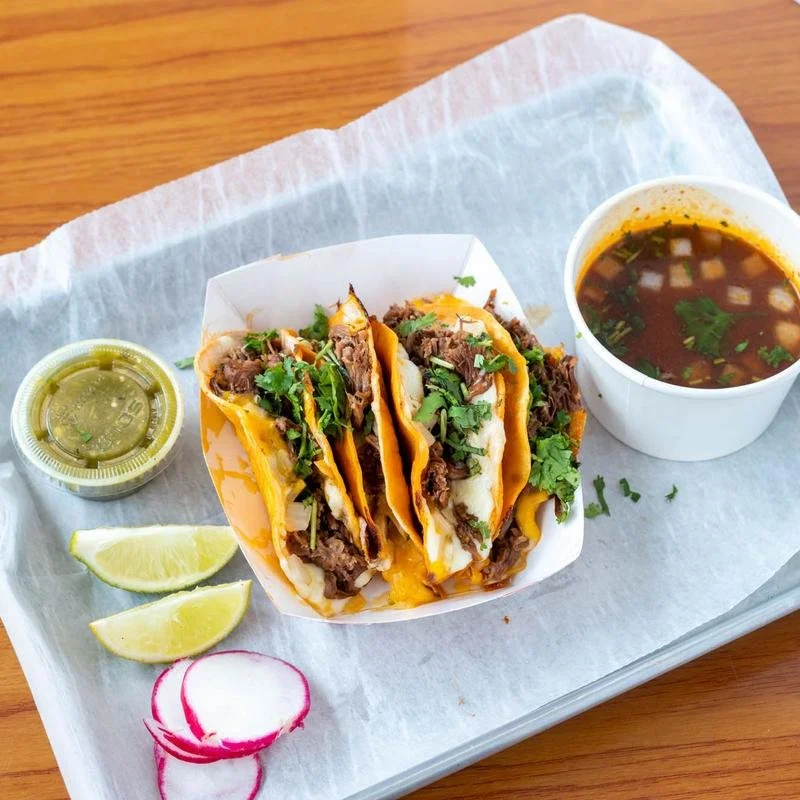 Three beef tacos topped with cilantro, served with lime wedges, sliced radish, and a side of cutting green salsa, along with a cup of beef broth with green onions and cilantro.