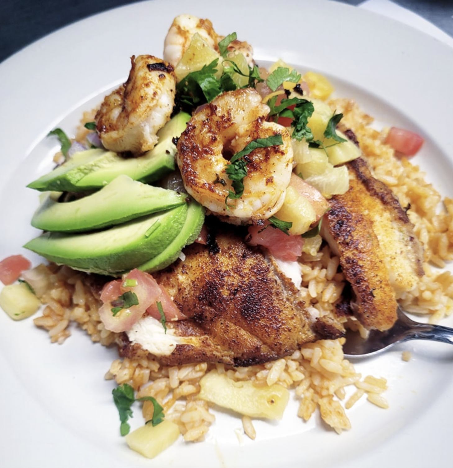 Plate of Mexican-style grilled chicken topped with shrimp, served over rice with avocado slices, pineapple chunks, tomatoes, and chopped herbs.