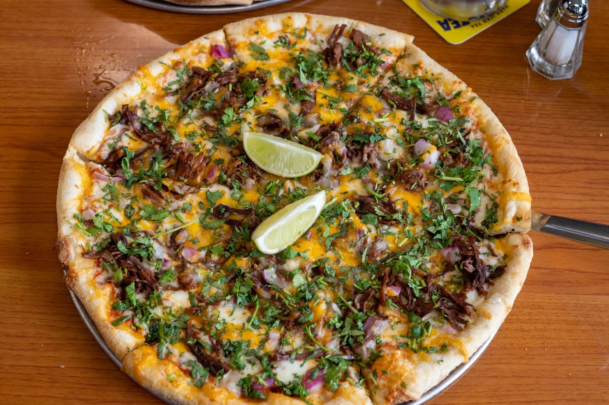 A pizza topped with shredded cheese, chopped cilantro, shredded beef, and lime wedges, on a wooden table with salt and pepper shakers nearby.