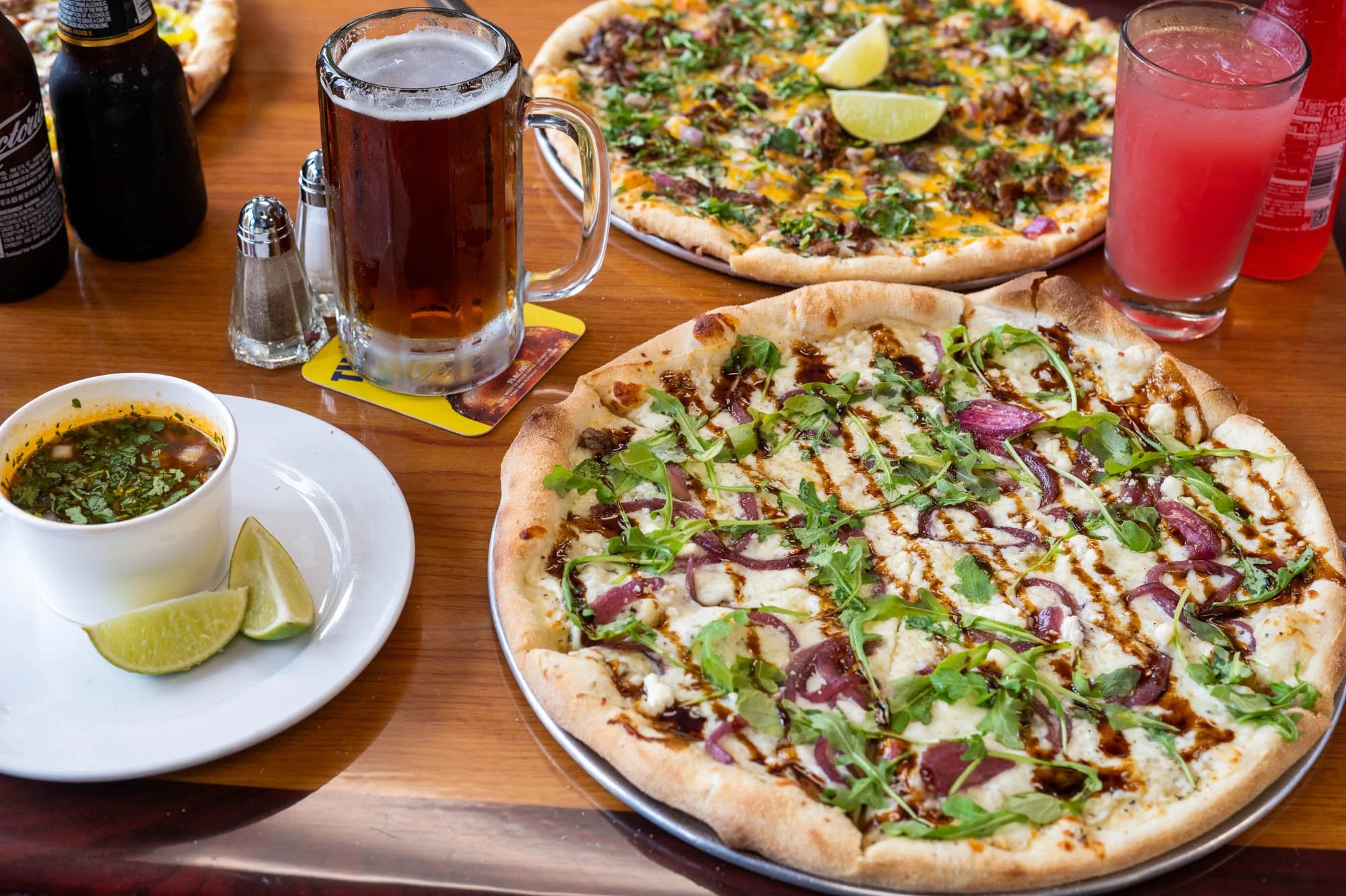 Two pizzas on a wooden table, one with toppings including arugula, onions, and a drizzle of balsamic glaze, the other with beef, cheese, cilantro, and lemon wedges, alongside a bowl of soup, a glass of dark soda, a pink drink, and condiment shakers.