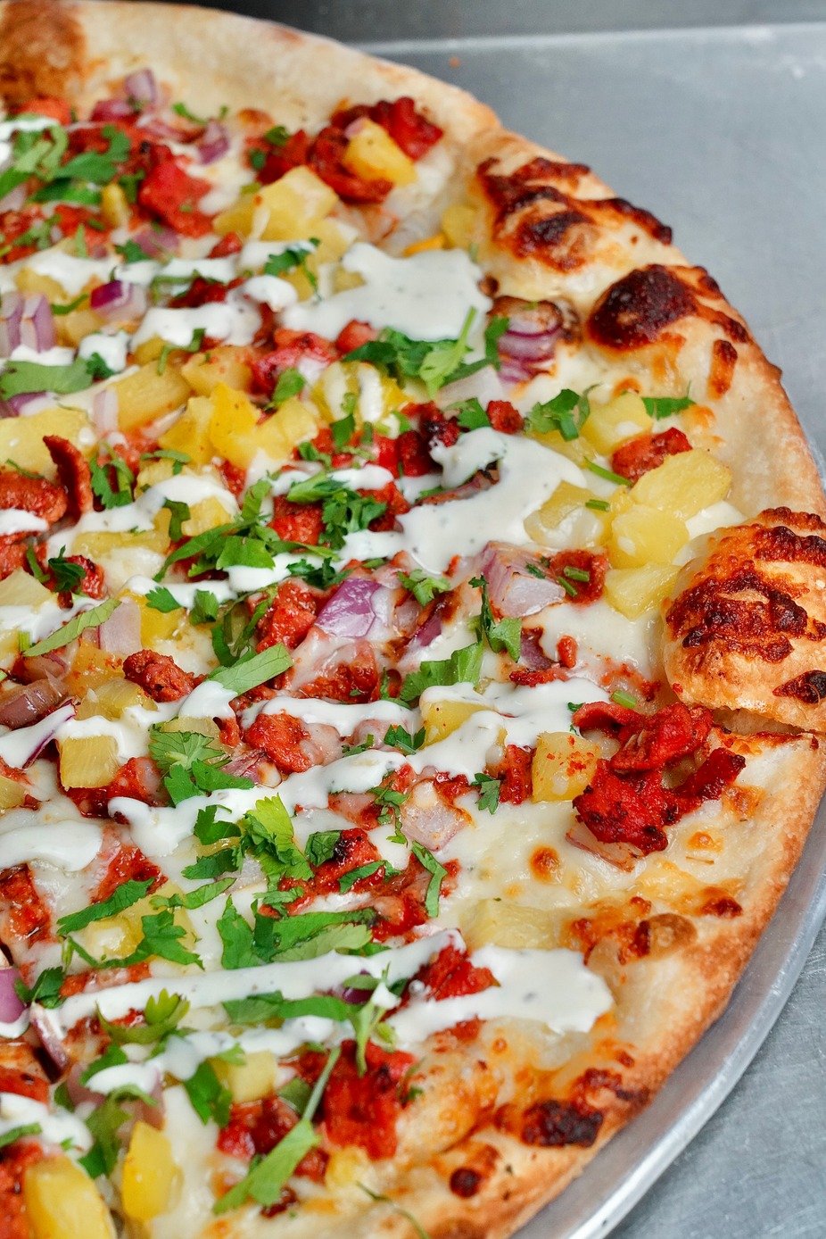 Close-up of a pizza with cheese, pineapple, red onions, green herbs, bacon pieces, and drizzled white sauce on a metal tray.