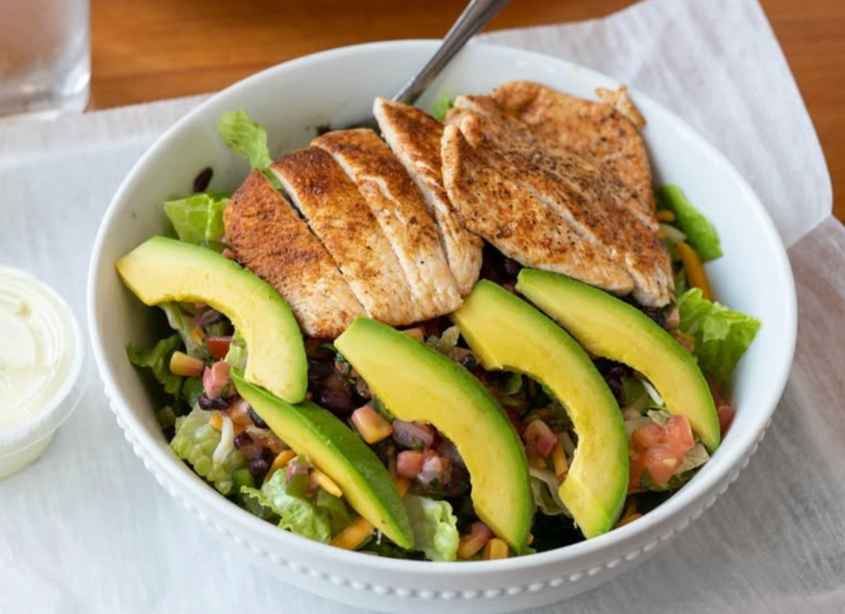 Salad bowl with grilled chicken, avocado slices, black beans, chopped tomatoes, lettuce, and shredded cheese.