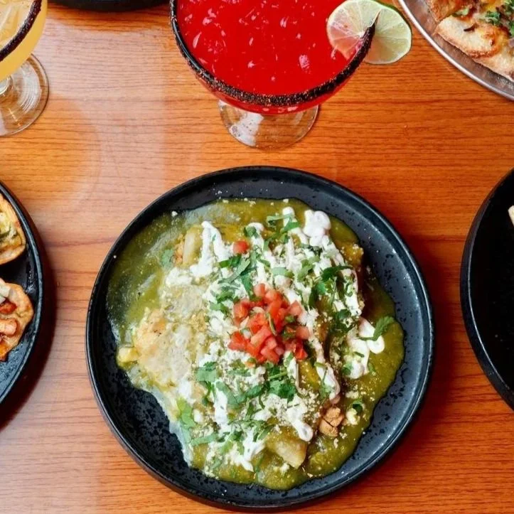 Plate of green chili with chopped tomatoes, chopped cilantro, shredded cheese, and chopped onions. Two cocktails with lime garnishes are partially visible at the top.