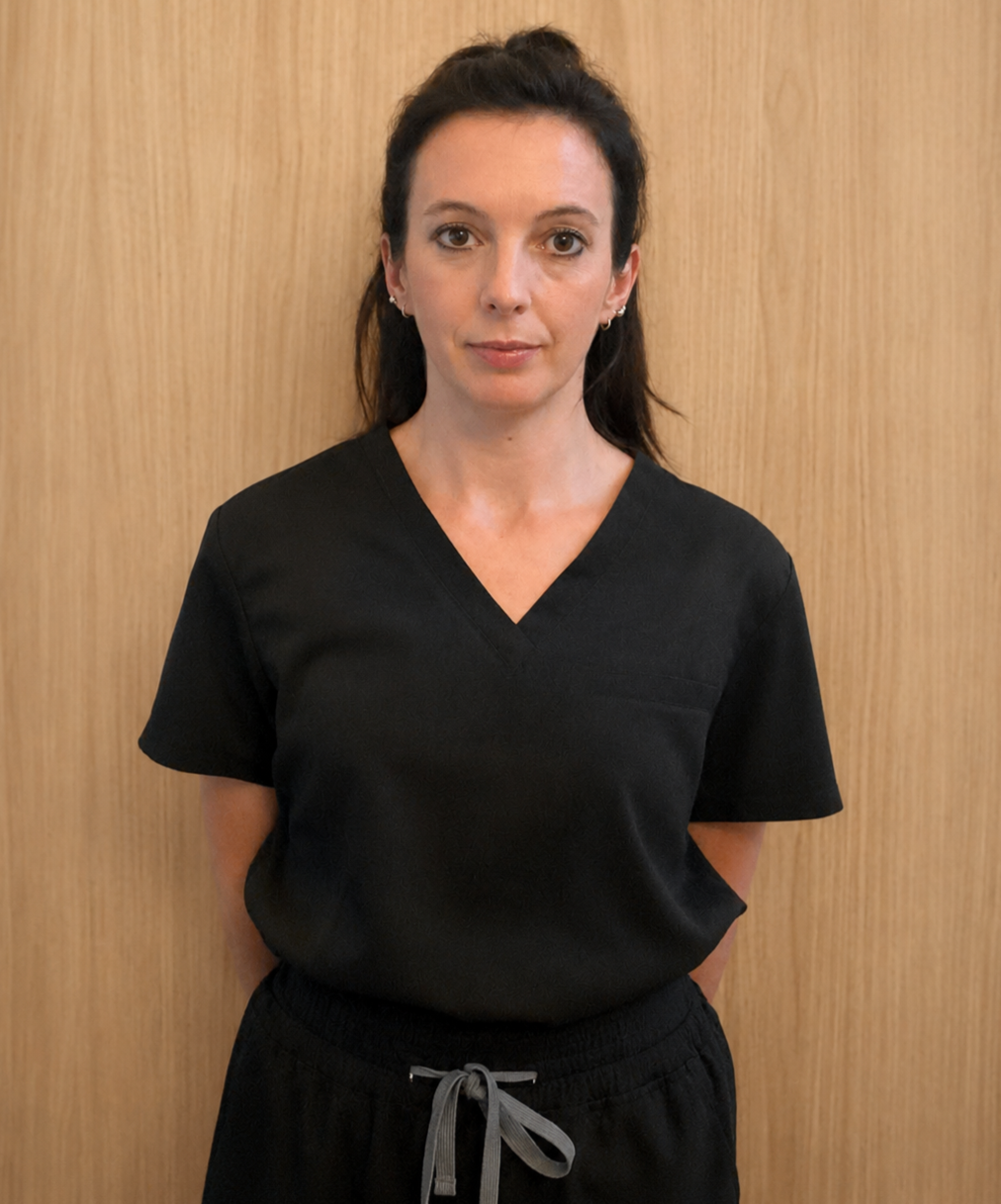 A woman with dark hair wearing black scrubs standing against a wood-paneled background.