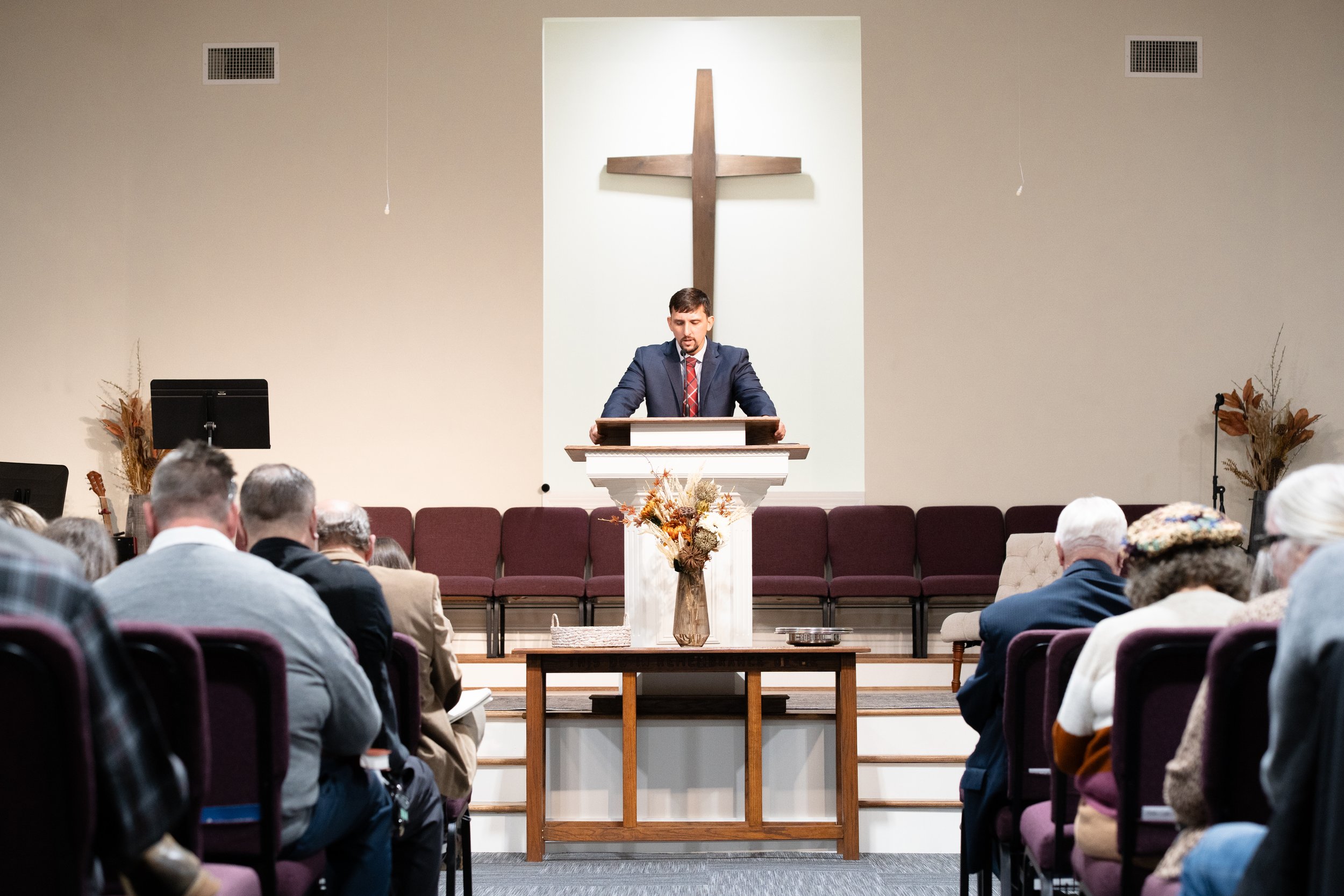Man in a suit and tie standing at a pulpit with a large wooden cross on the wall behind him, addressing a congregation in a church or chapel.
